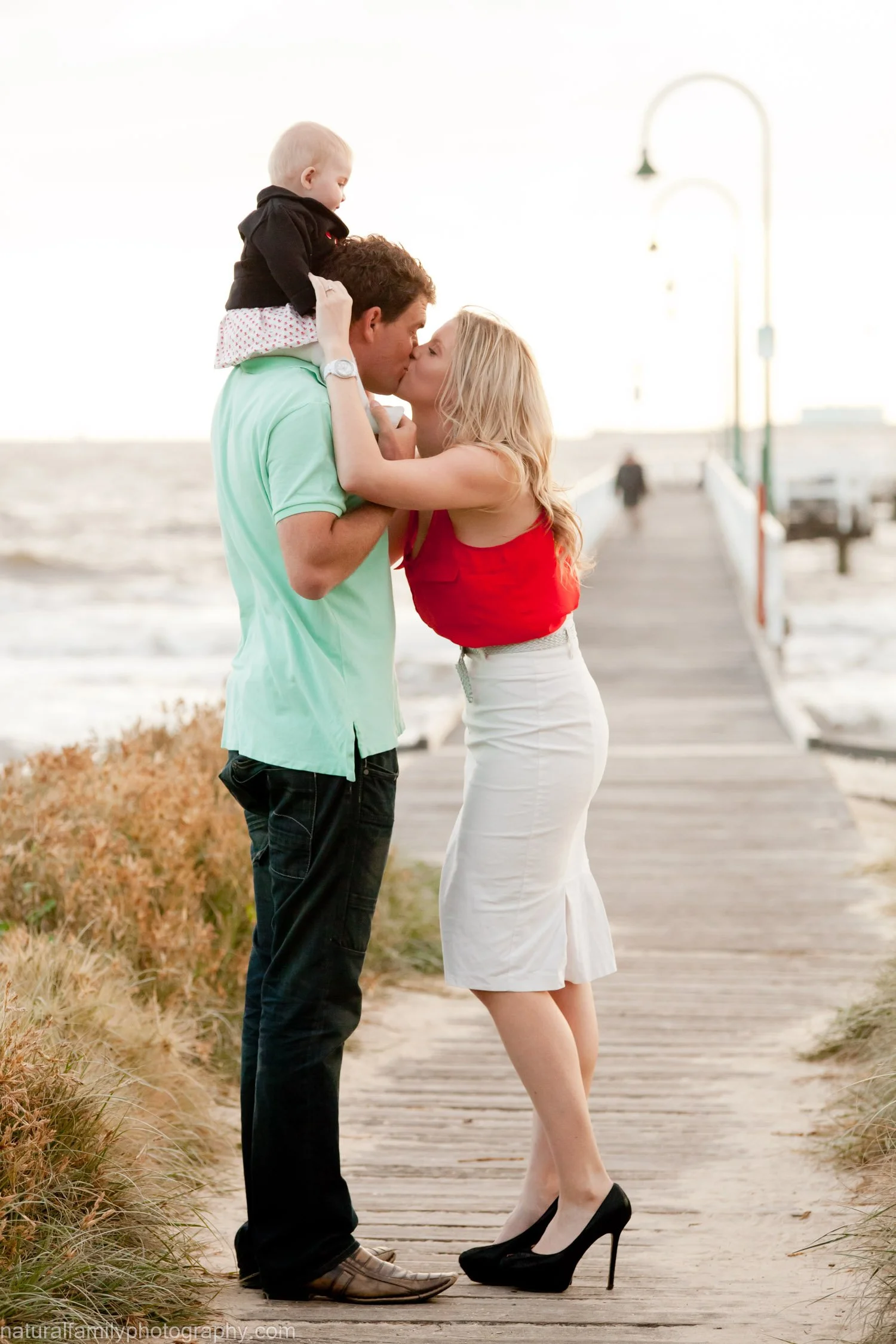 A couple sharing a kiss on a dock by the water, with a young child sitting on the man's shoulders, during sunset.