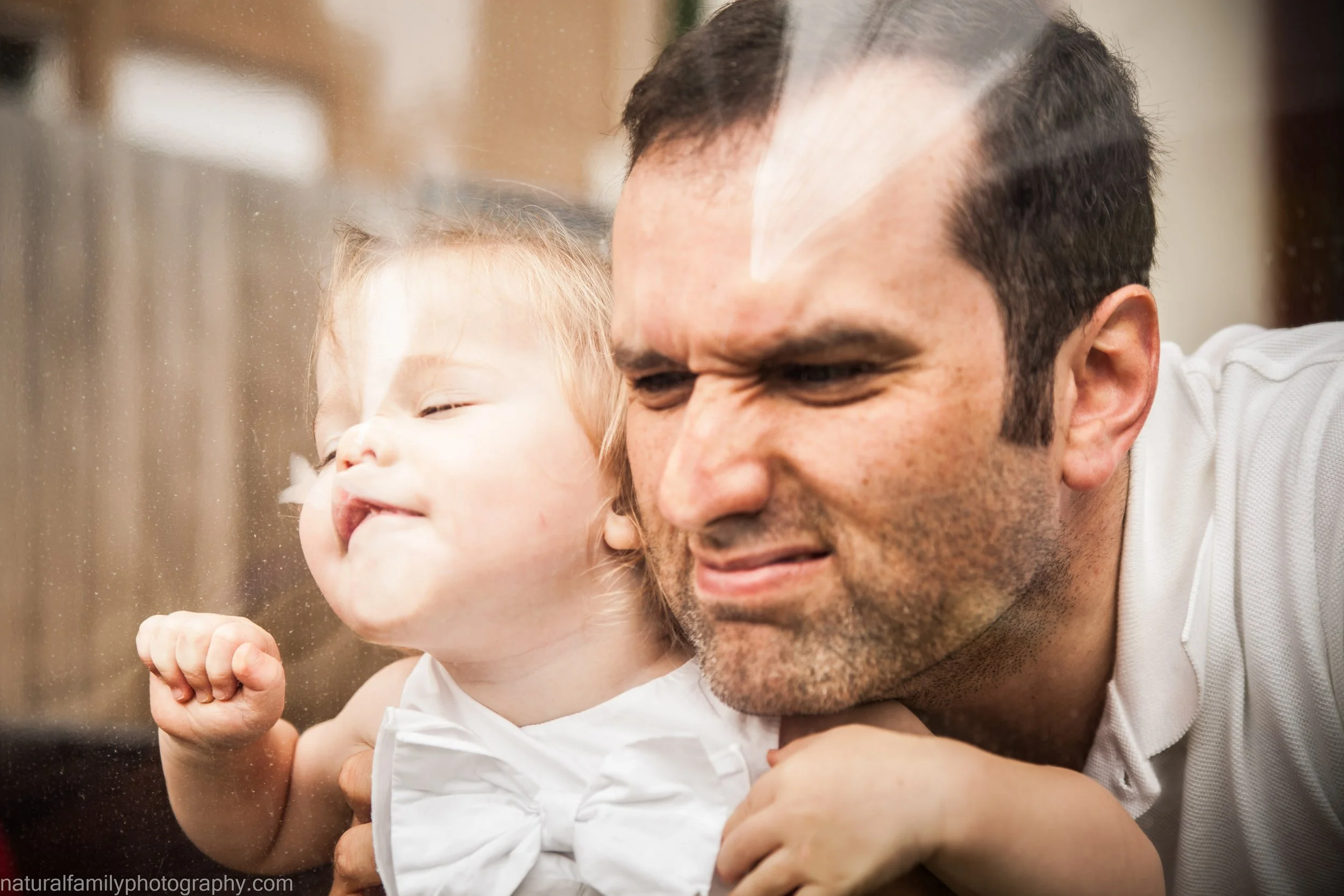 A man and a young girl pressing their faces against a glass window, making funny faces. The girl has her eyes closed and an expression of amusement, while the man looks slightly squished, both enjoying a playful moment.