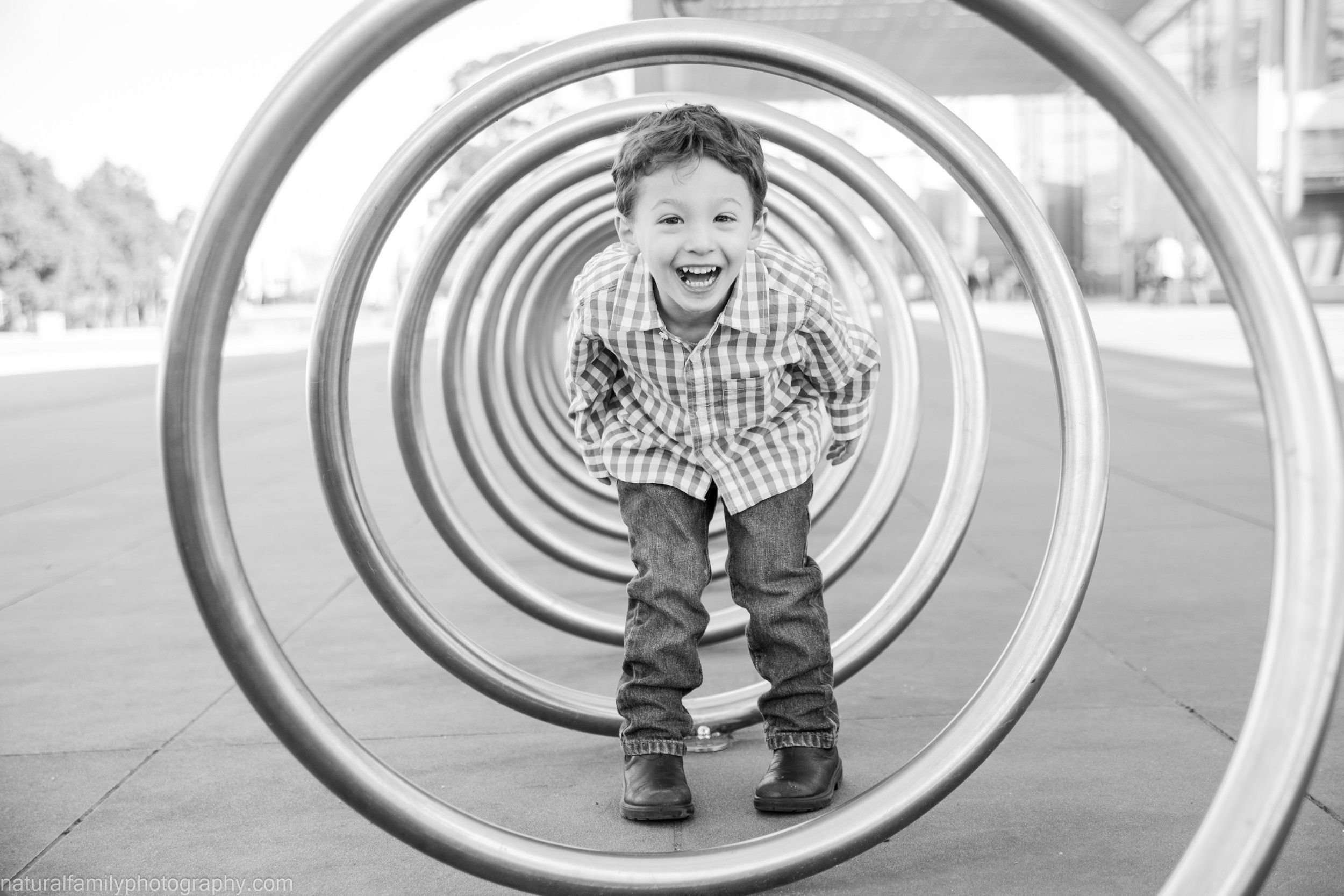 A young boy smiling and crawling through a circular metal tunnel at a playground, black and white photo.