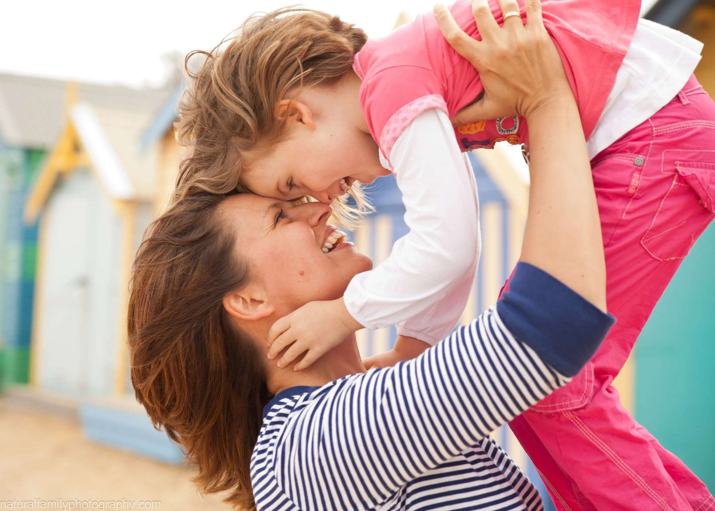 A woman lifting a young girl in the air, both smiling and touching foreheads, outdoors near a colorful playground.
