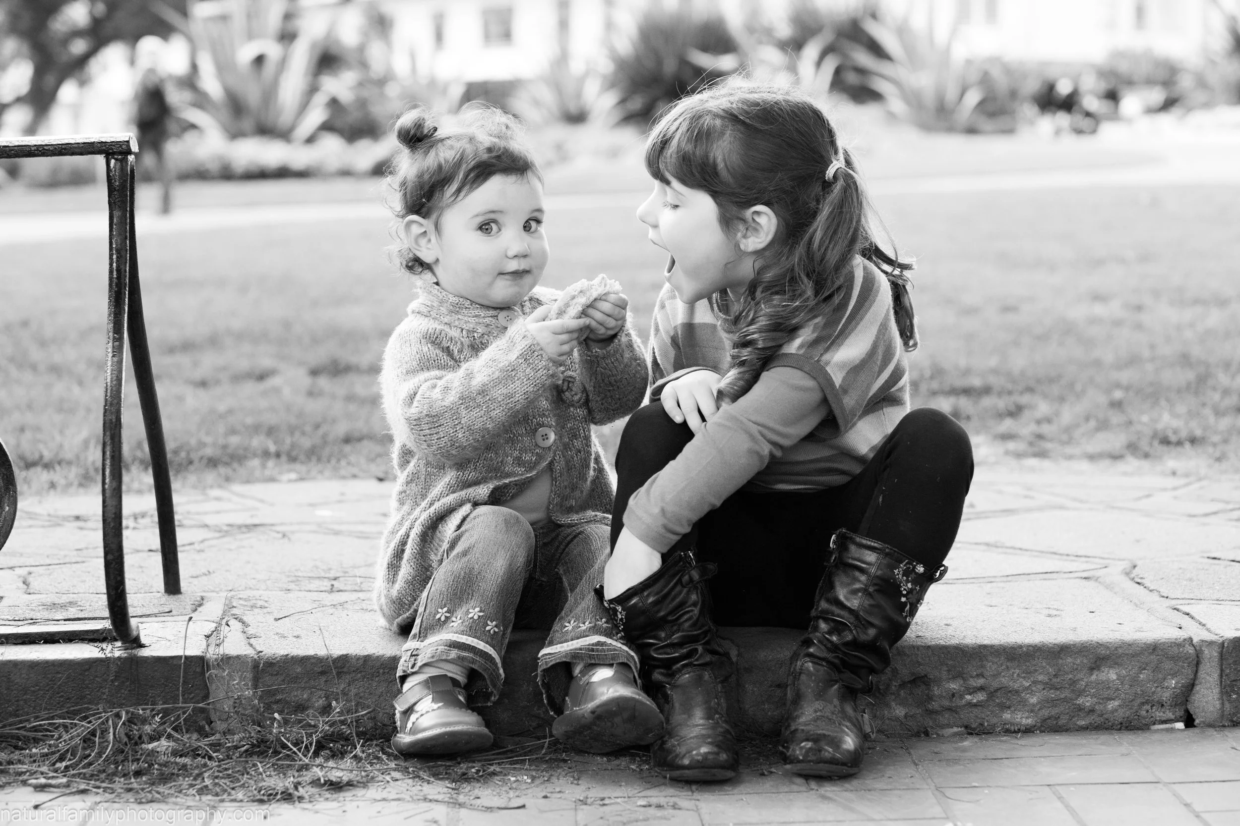 A young girl sitting on a curb and a toddler girl, with the older girl animatedly talking to the younger girl who is holding a snack, in a park or outdoor setting.