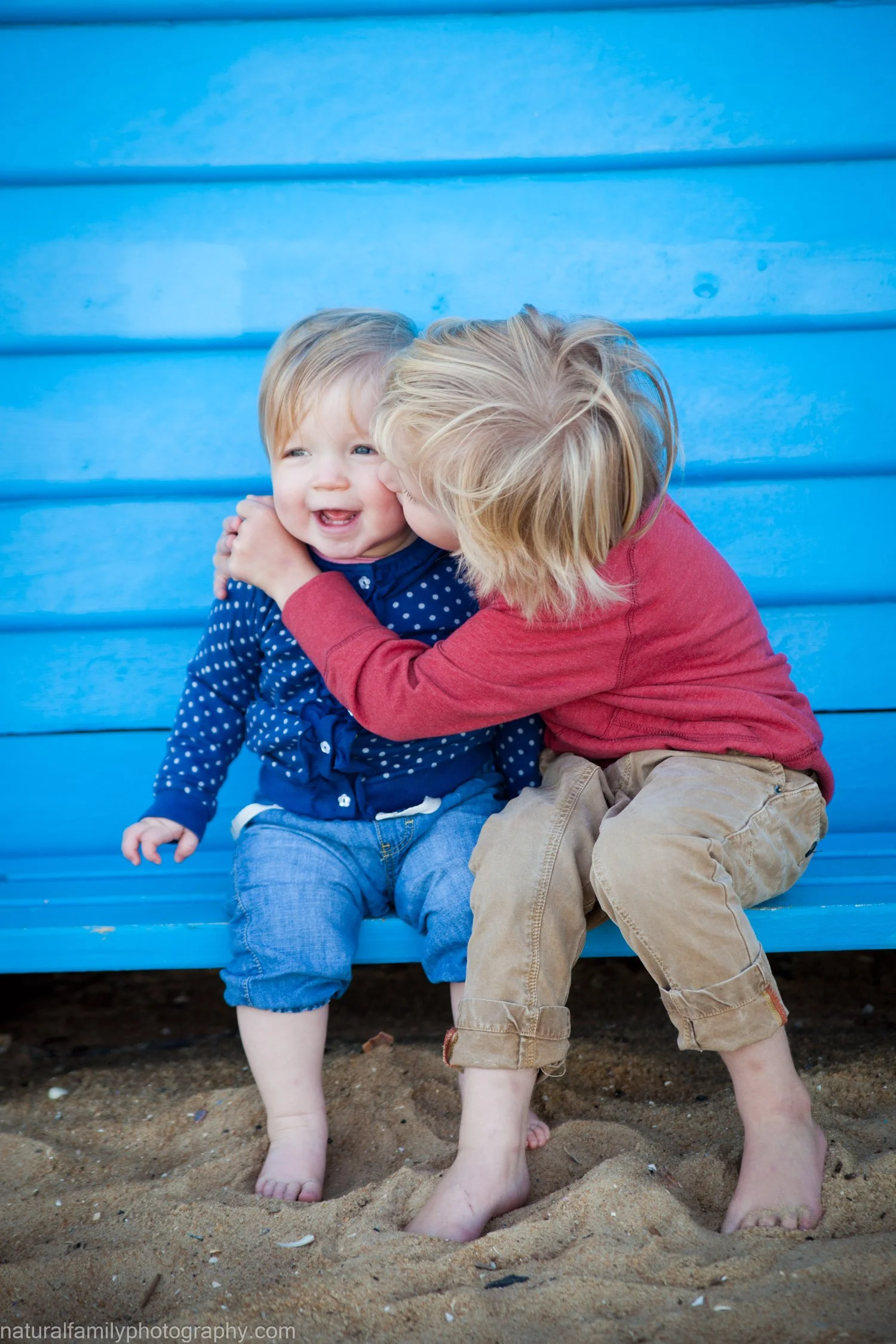 Two young children, a boy and a girl, sitting on a blue bench at the beach, sharing a joyful moment with the girl giving the boy a kiss on the cheek.