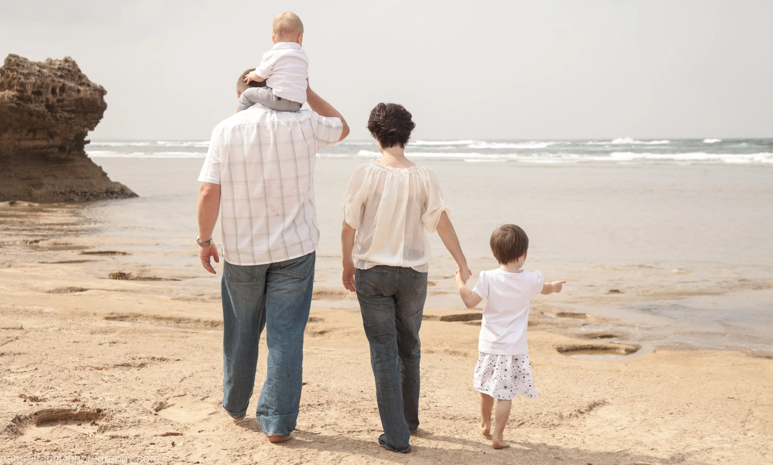 Family of four walking on the beach, holding hands, with a rocky formation and the ocean in the background.