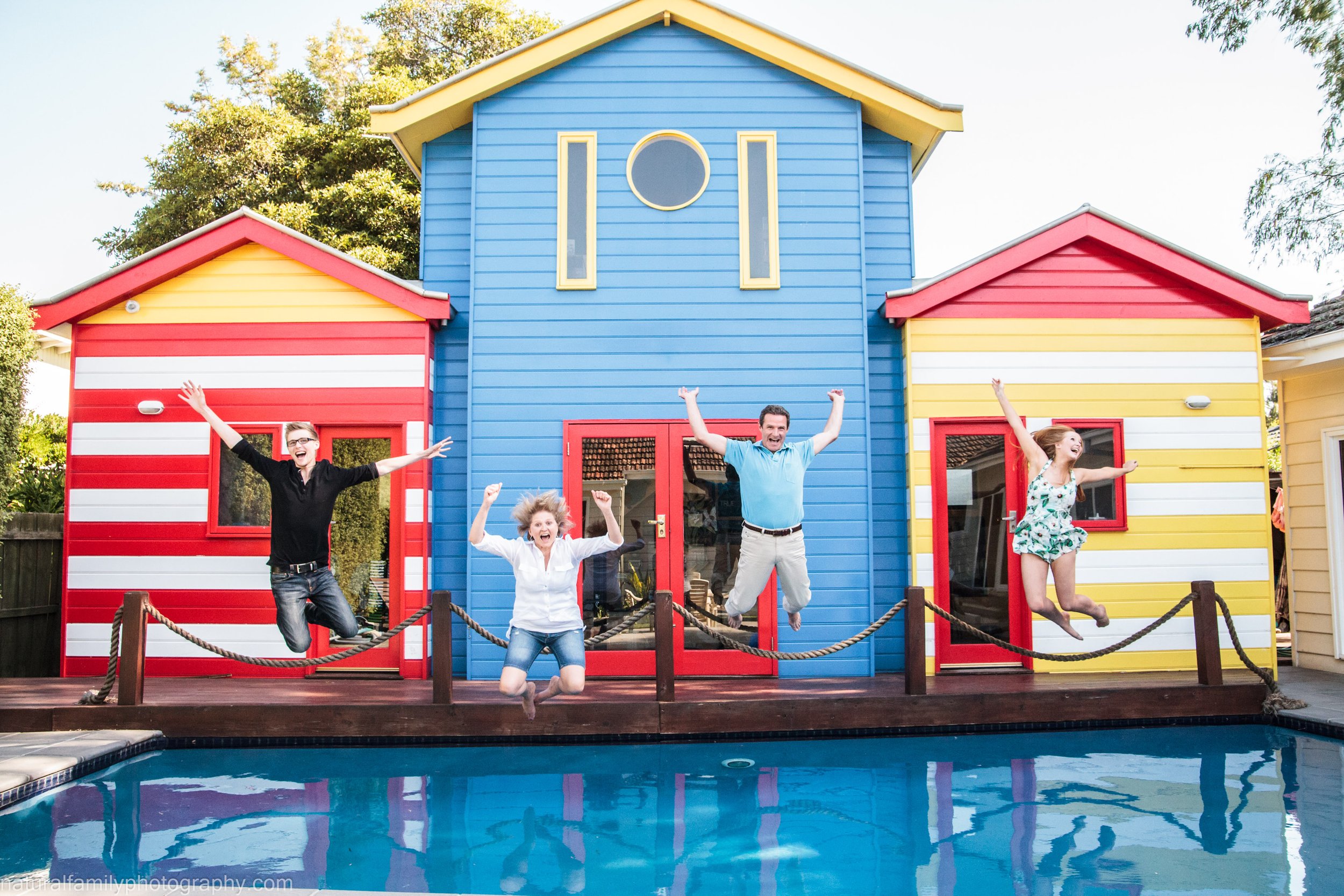 Four people jumping on a wooden deck in front of colorful beach houses, with a swimming pool in the foreground.