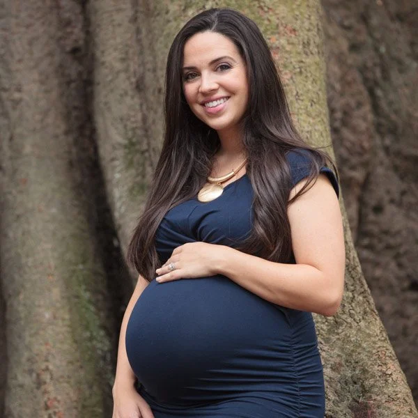 Pregnant woman with long dark hair smiling and standing outdoors near a large tree, wearing a navy blue dress. Maternity portraiture by Natural Family Photography, Melbourne.