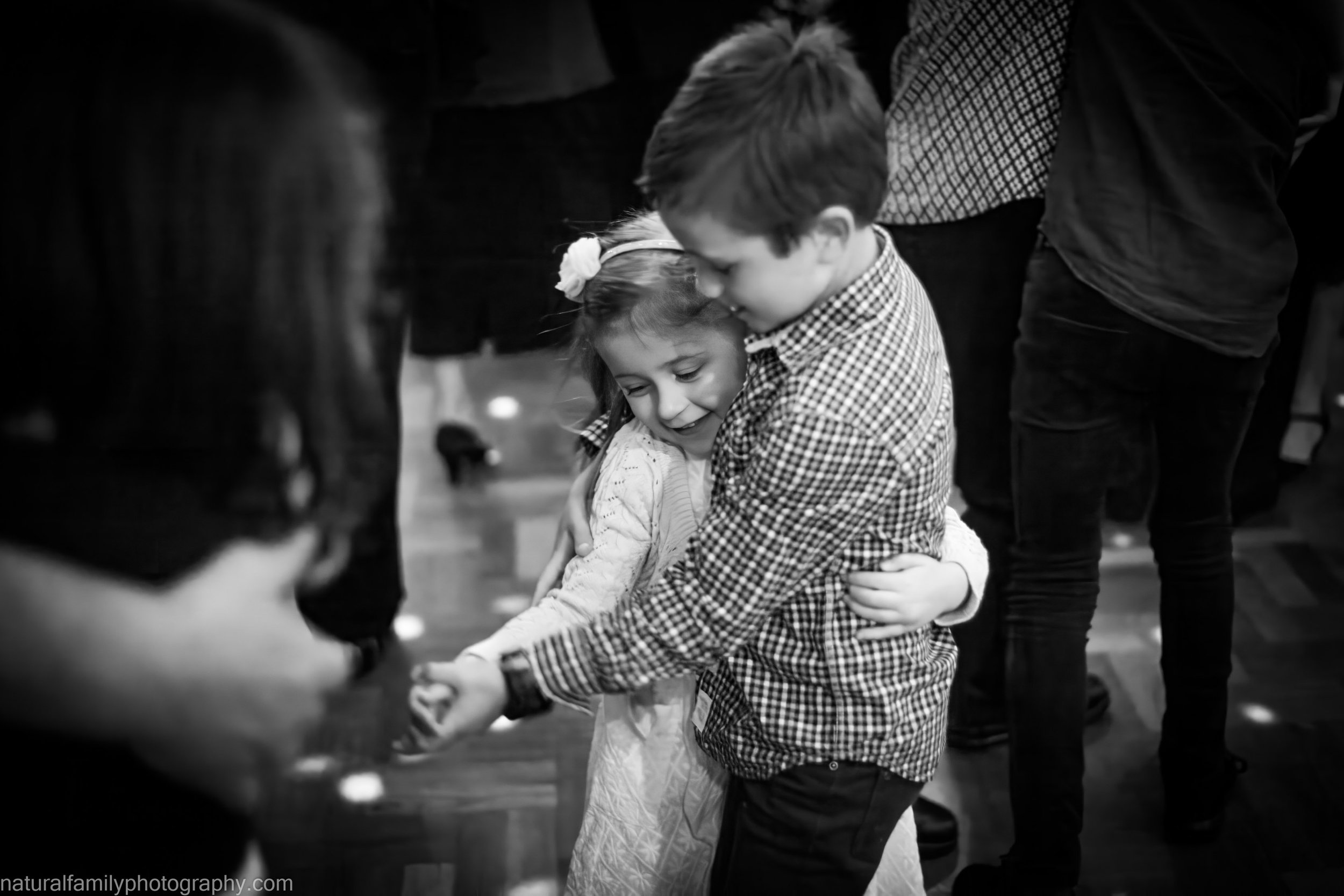 A young boy and girl share an embrace and smile at each other during a dance, surrounded by adults. Classic black and white portrait by Natural Family Photography, Melbourne.