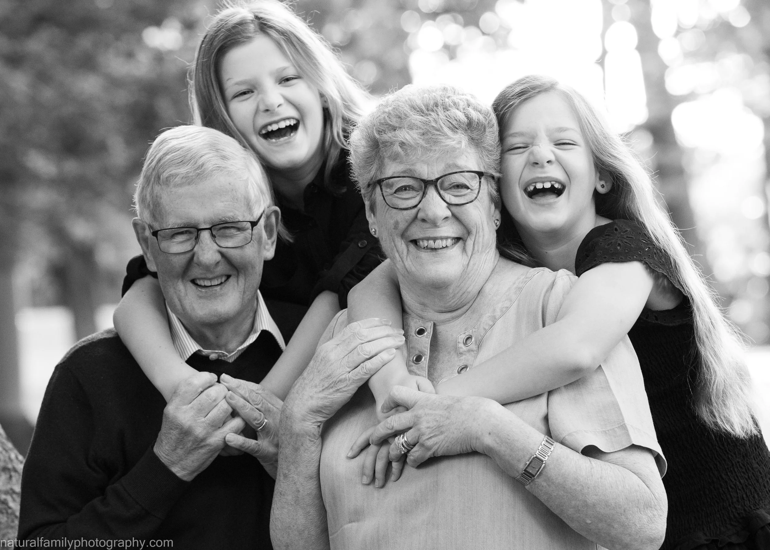 A joyful multigeneration family consisting of two elderly grandparents, a grandfather and grandmother, with two young girls, smiling and hugging outdoors.