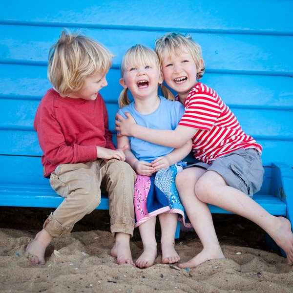 Three young children sitting on a sandy beach, laughing and hugging each other in front of a bright blue wooden wall. Brighton beach box family portraits by Natural Family Photography, Melbourne.