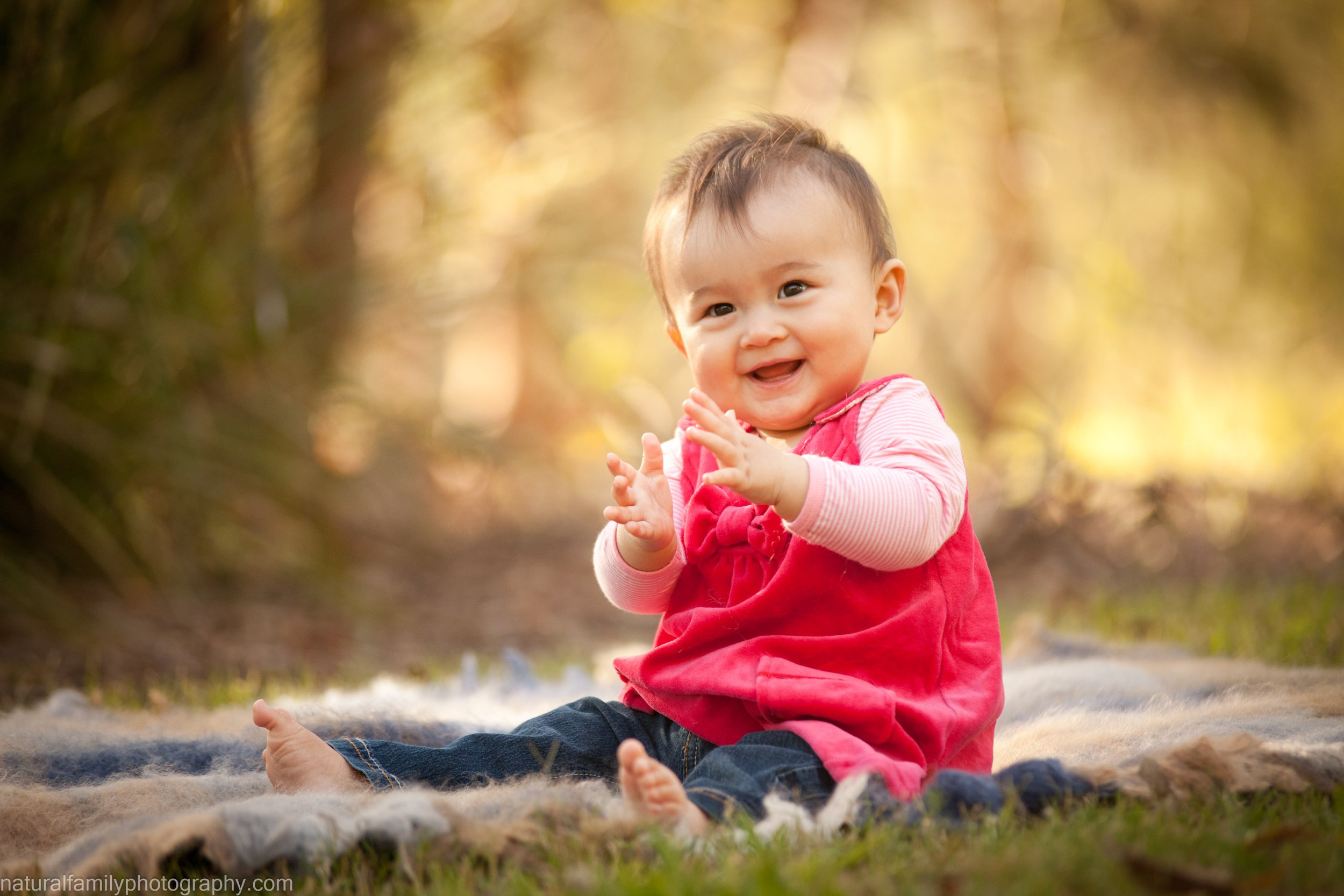 A smiling baby girl sitting on a blanket outdoors in a wooded area during golden hour, wearing a pink dress and jeans.