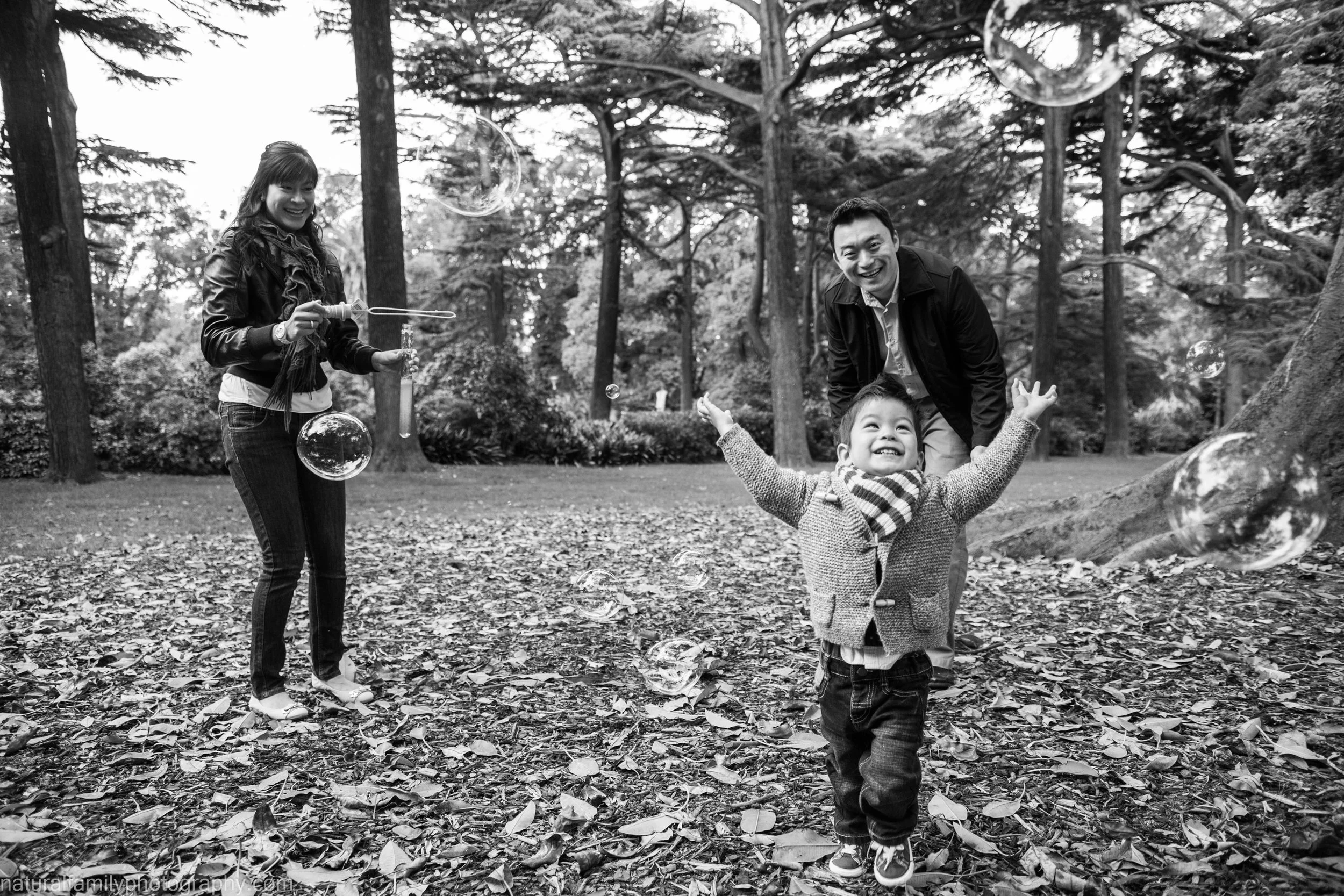 A family enjoying a playful moment outdoors in a park, with a woman blowing bubbles, a young boy with arms raised, and a man smiling behind the boy, surrounded by trees and fallen leaves.