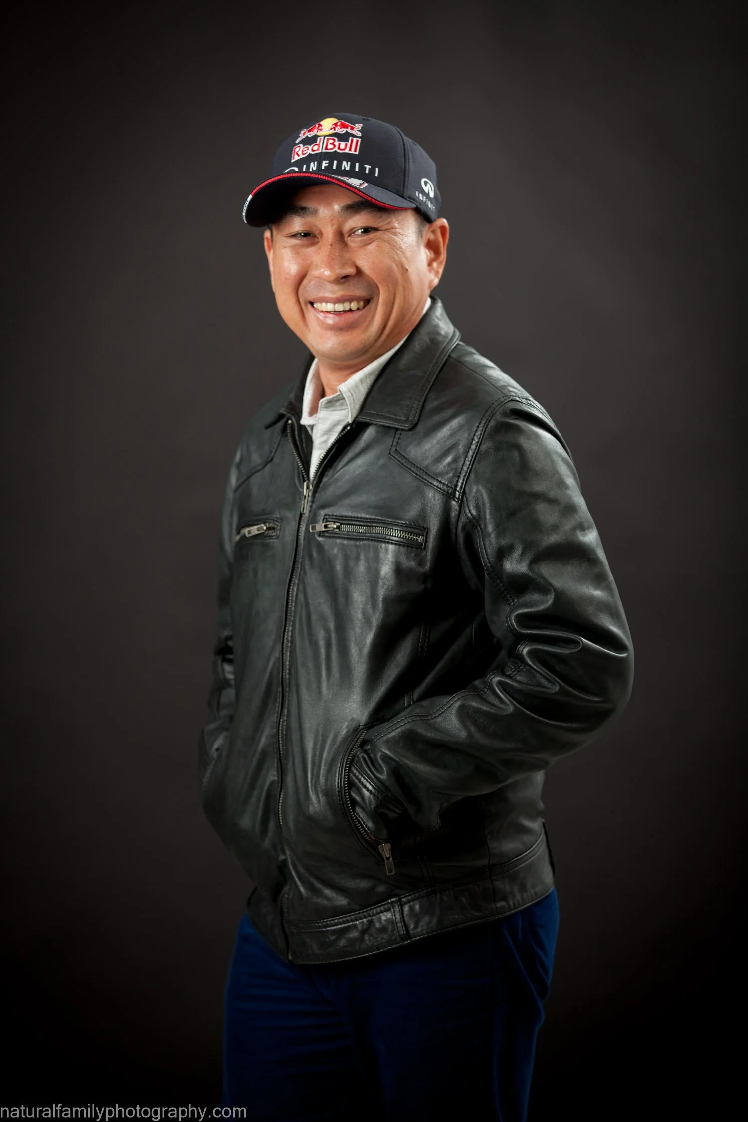 A smiling man in a black leather jacket, white shirt, and a Red Bull Racing cap against a dark background.