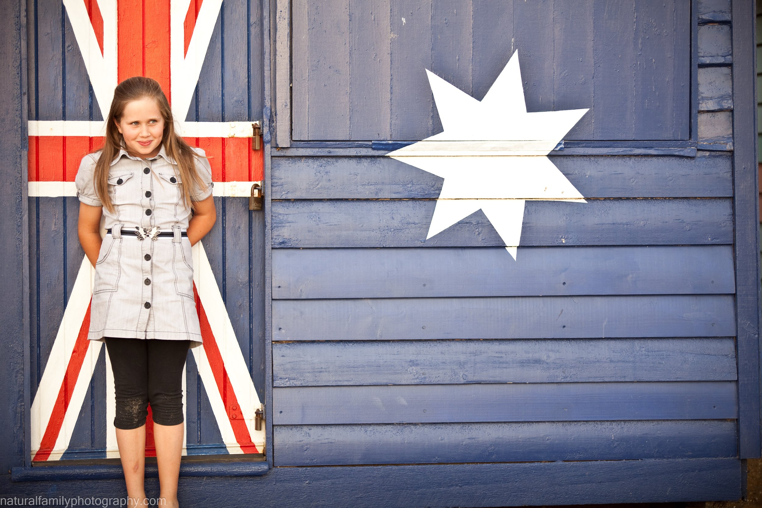Young girl standing beside a Brighton Beach Box with the Australian flag painted on it. Portrait by Natural Family Photography, Melbourne.