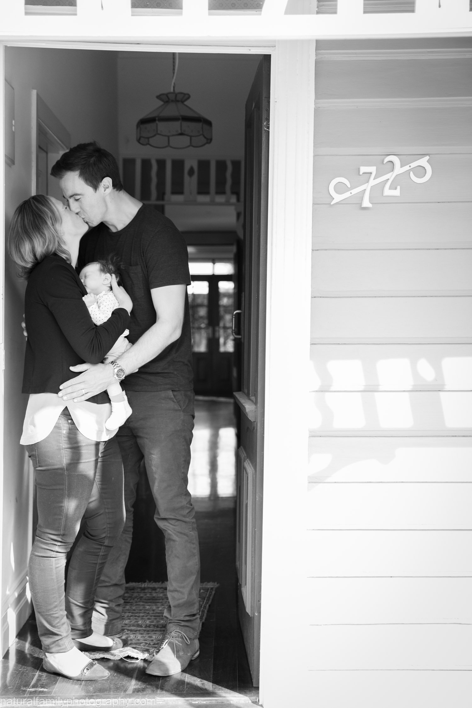 A family sharing a kiss at their home doorway, holding a newborn baby.