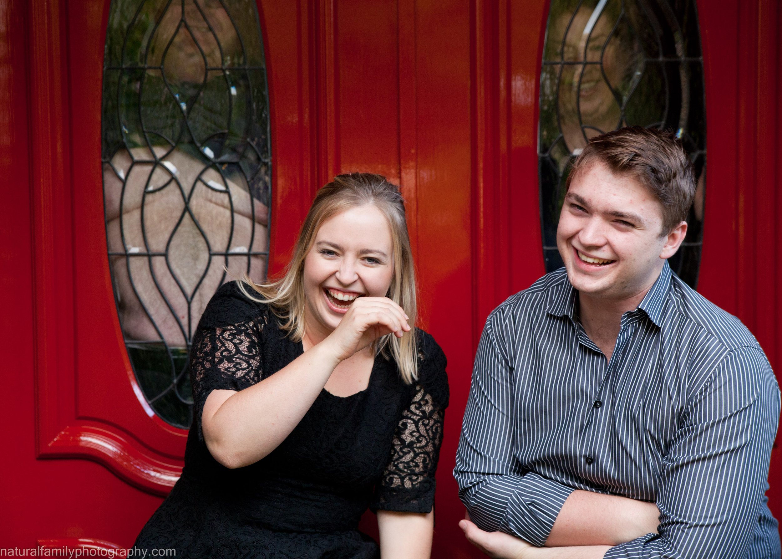 Two young adults, a woman and a man, are standing in front of a red door with decorative glass windows. They are smiling and laughing because their father is pressing his bare chest against the glass.