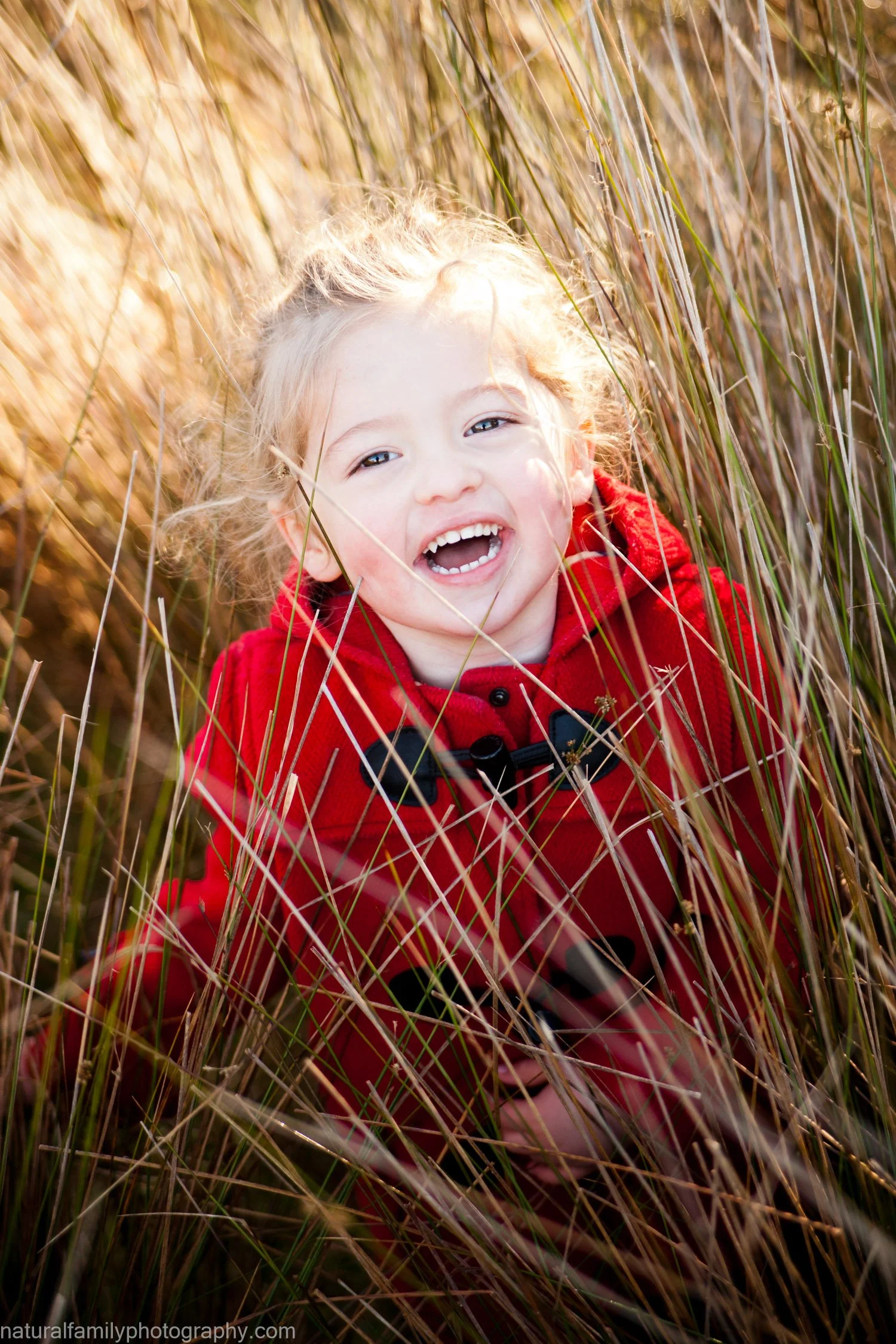 A young girl with blond hair smiling and laughing while crawling through tall dry grass, wearing a red jacket.