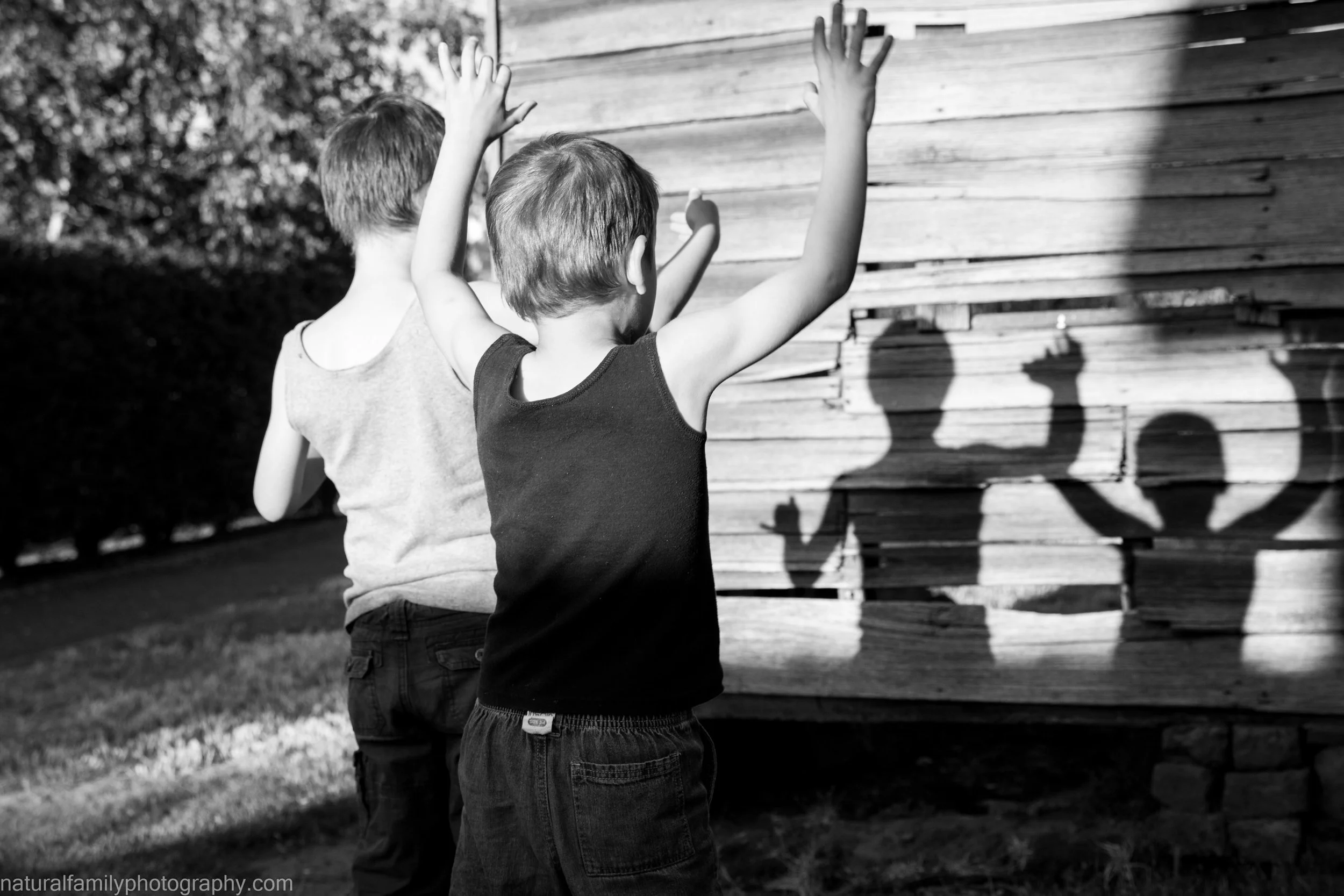 Two children, a boy wearing a black tank top and another child in a sleeveless shirt, are seen with their backs to the camera, throwing shadow puppets on a wooden wall with the sunlight.