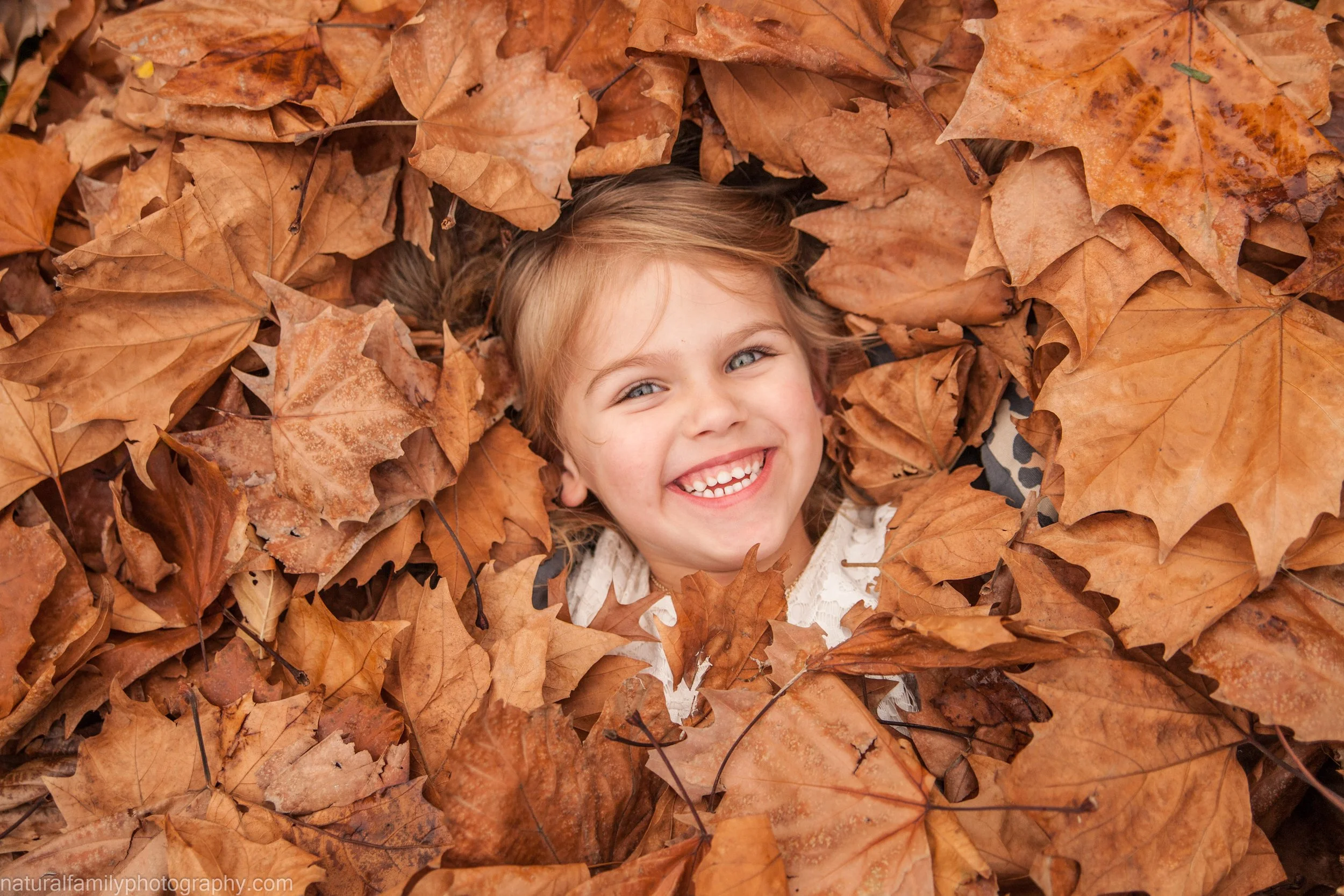 A young girl with blonde hair and a big smile lying in a pile of brown autumn leaves.