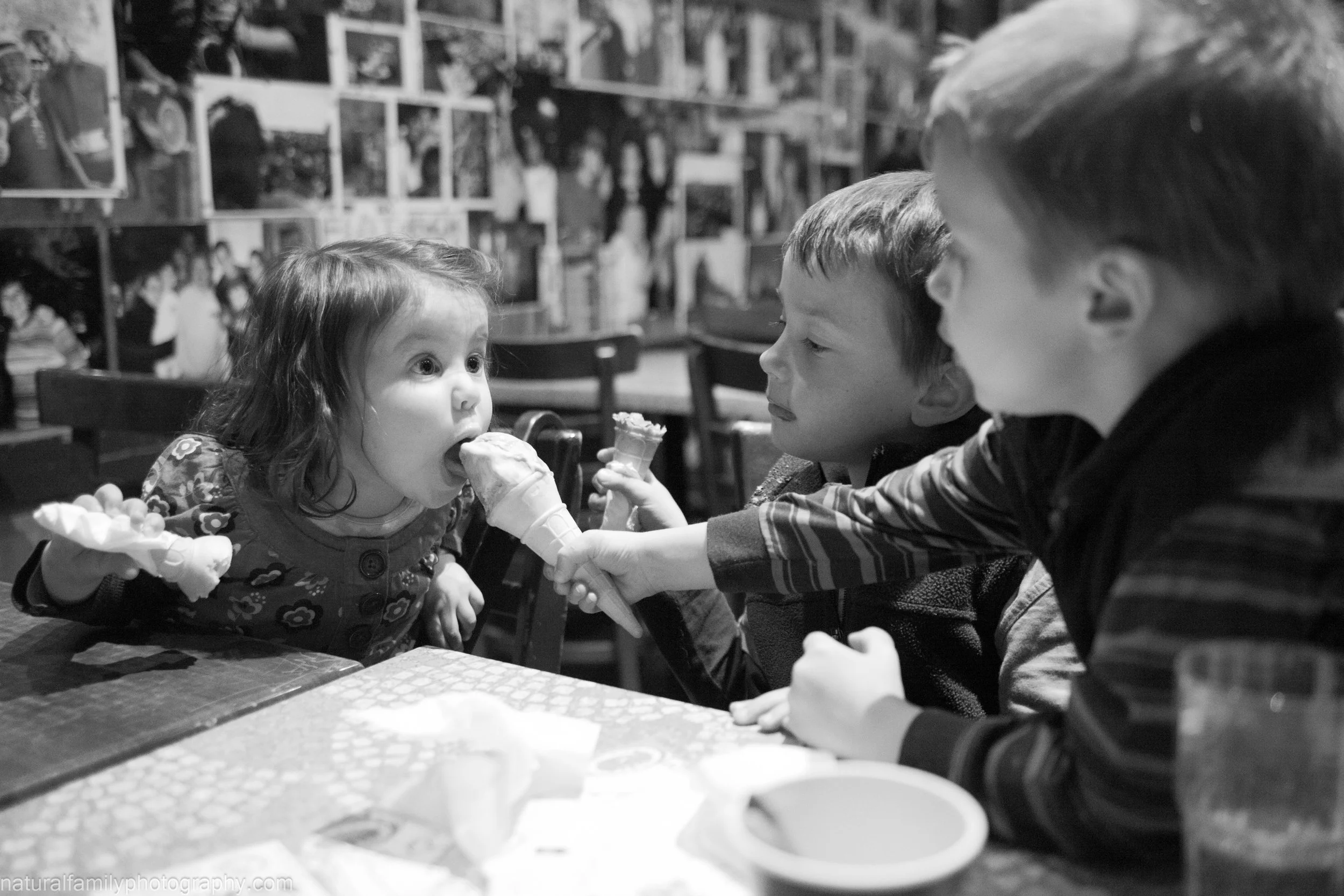 Three children sharing ice cream at a restaurant or cafe, with one girl taking a lick and another boy holding a cone.