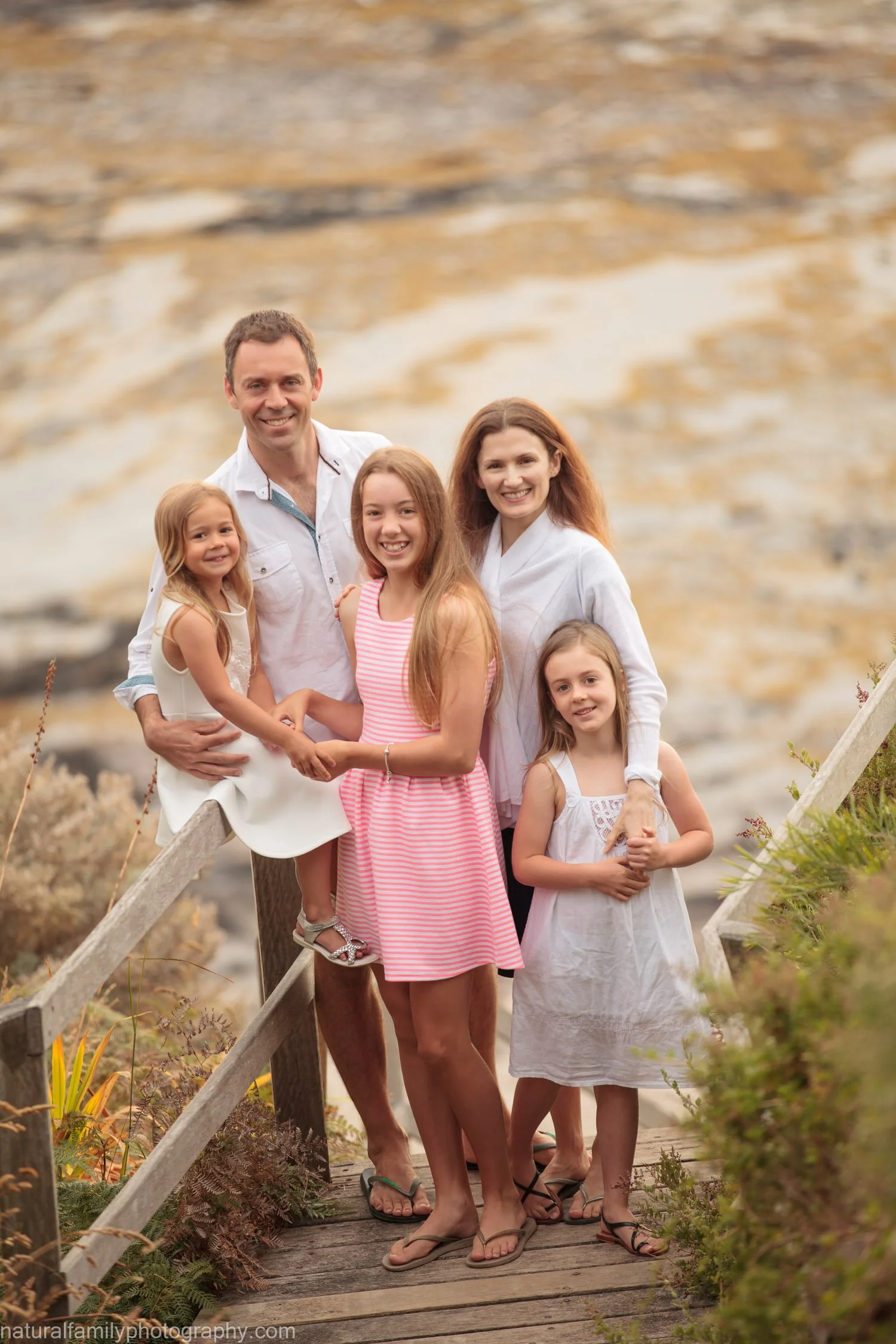A family of six, consisting of a father, mother, and four daughters, stands on a wooden walkway outdoors with a rocky cliff in the background. They are smiling and dressed in casual summer clothing.