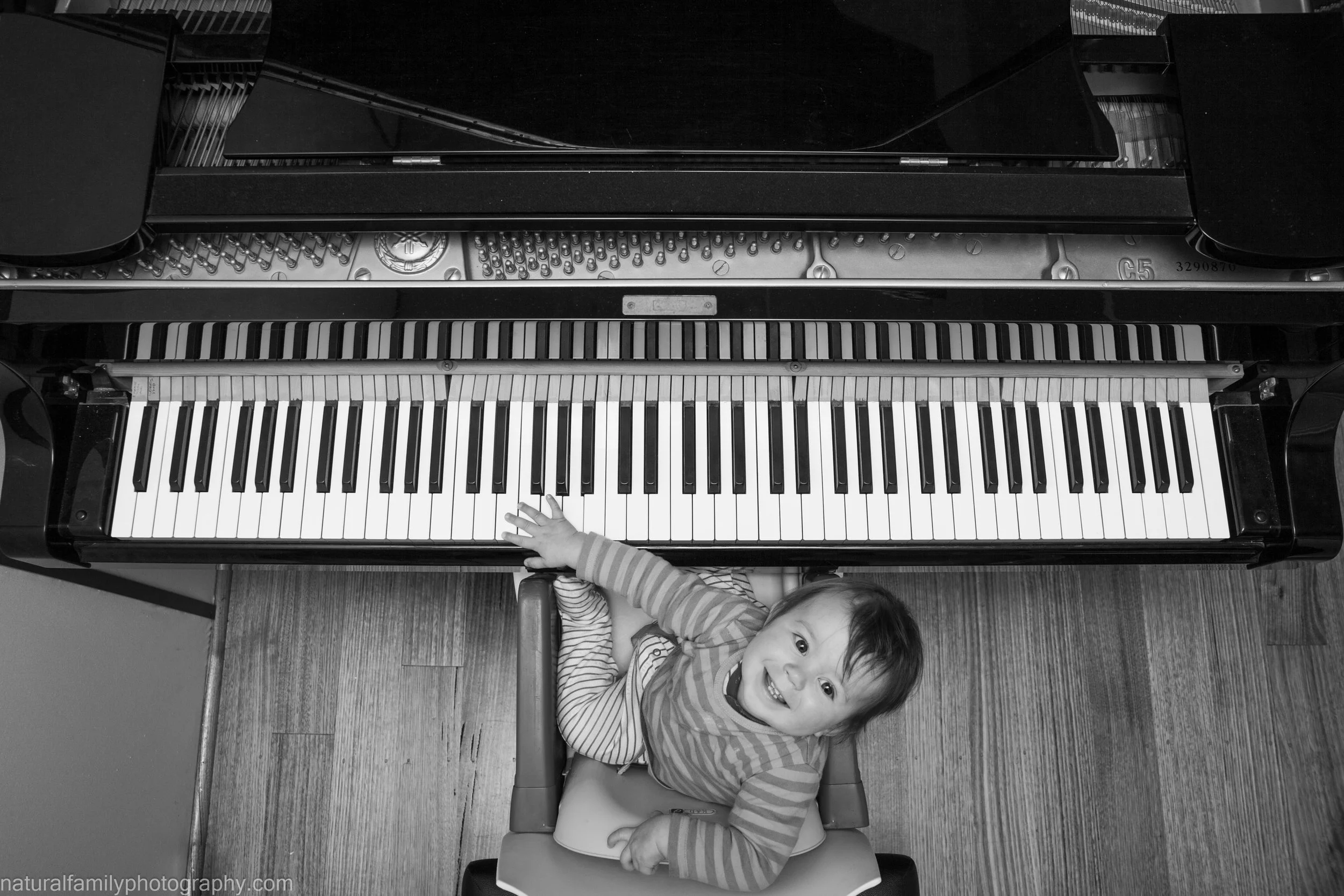 Child smiling and reaching towards the keys of a grand piano while sitting on a chair, viewed from above in black and white.