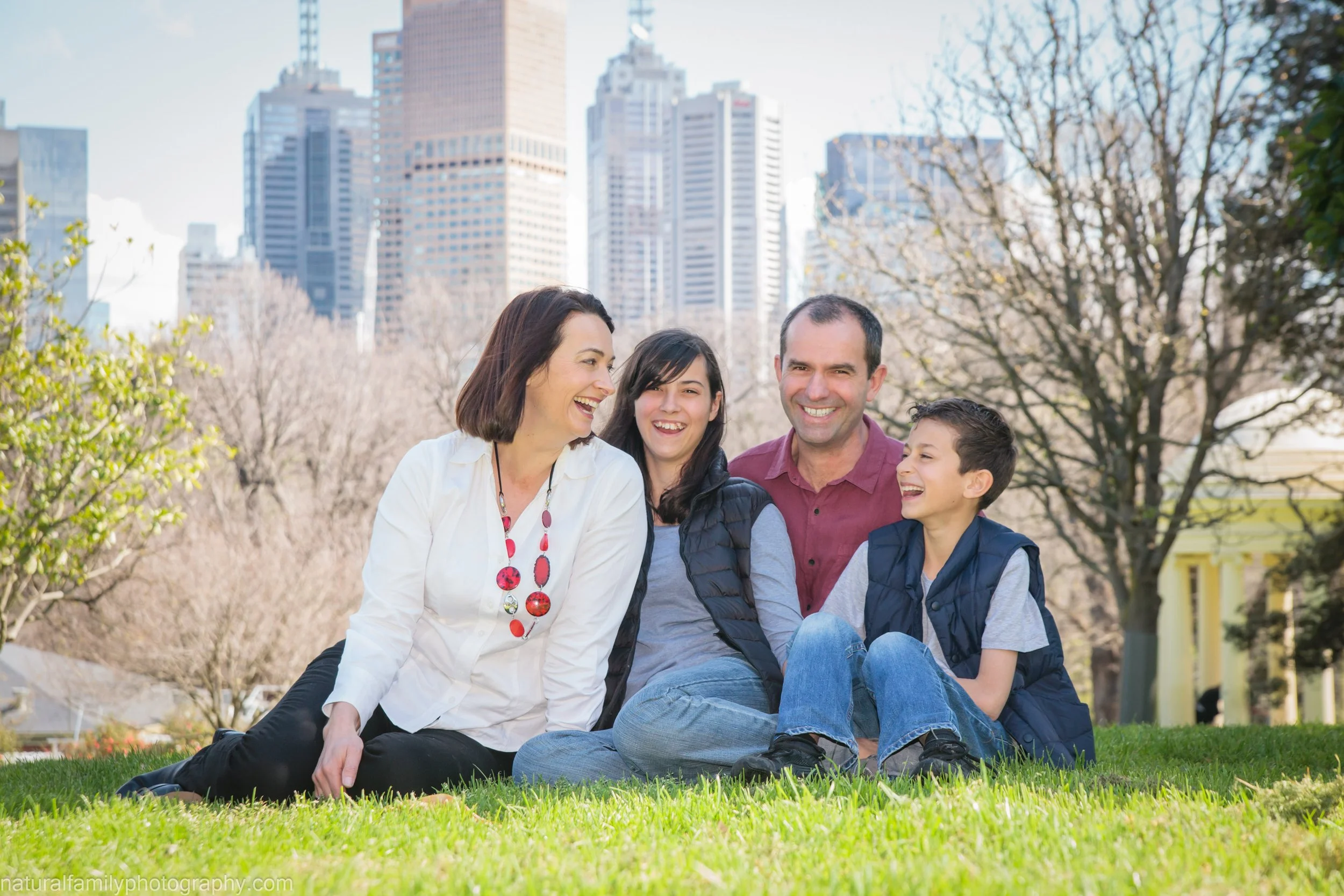 Family of four sitting on grass in a park, smiling and laughing, with city buildings in the background.