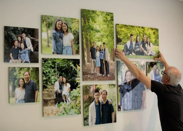 A professional artwork installer arranging clear-mounted family photos on a living room wall, featuring various outdoor portraits of a family in different natural settings. Custom-designed and produced by Natural Family Photography.