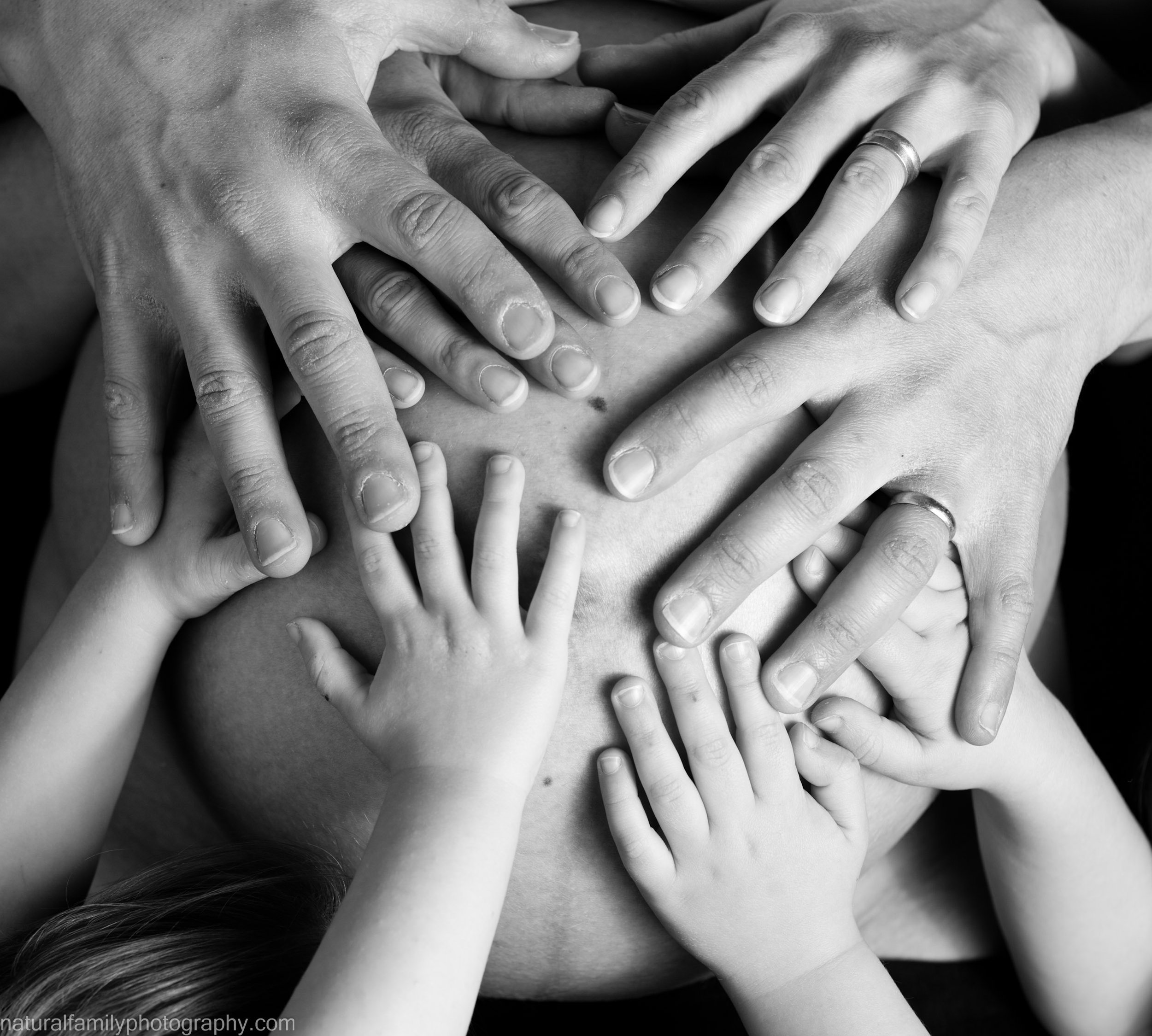 Multiple hands of various sizes, including small children's hands and adult hands, are placed on a mother's pregnant belly, creating a layered and intimate composition. Artistic black and white maternity photography by Natural Family Photography.