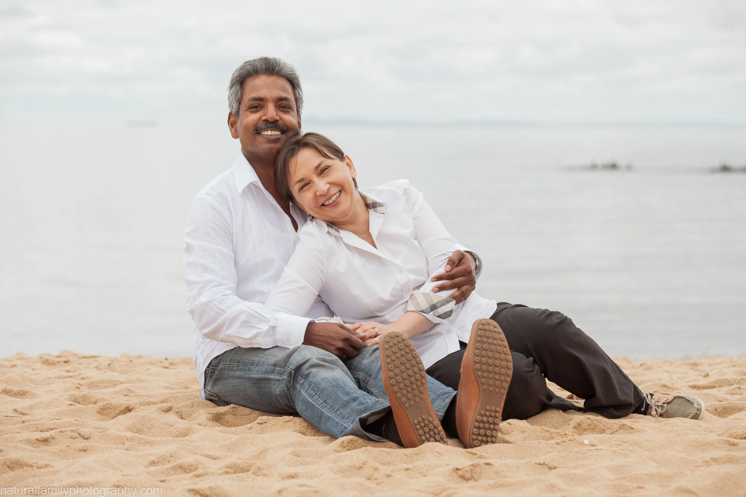 A happy couple sitting on the sand at the beach, smiling and embracing with trees and water in the background.