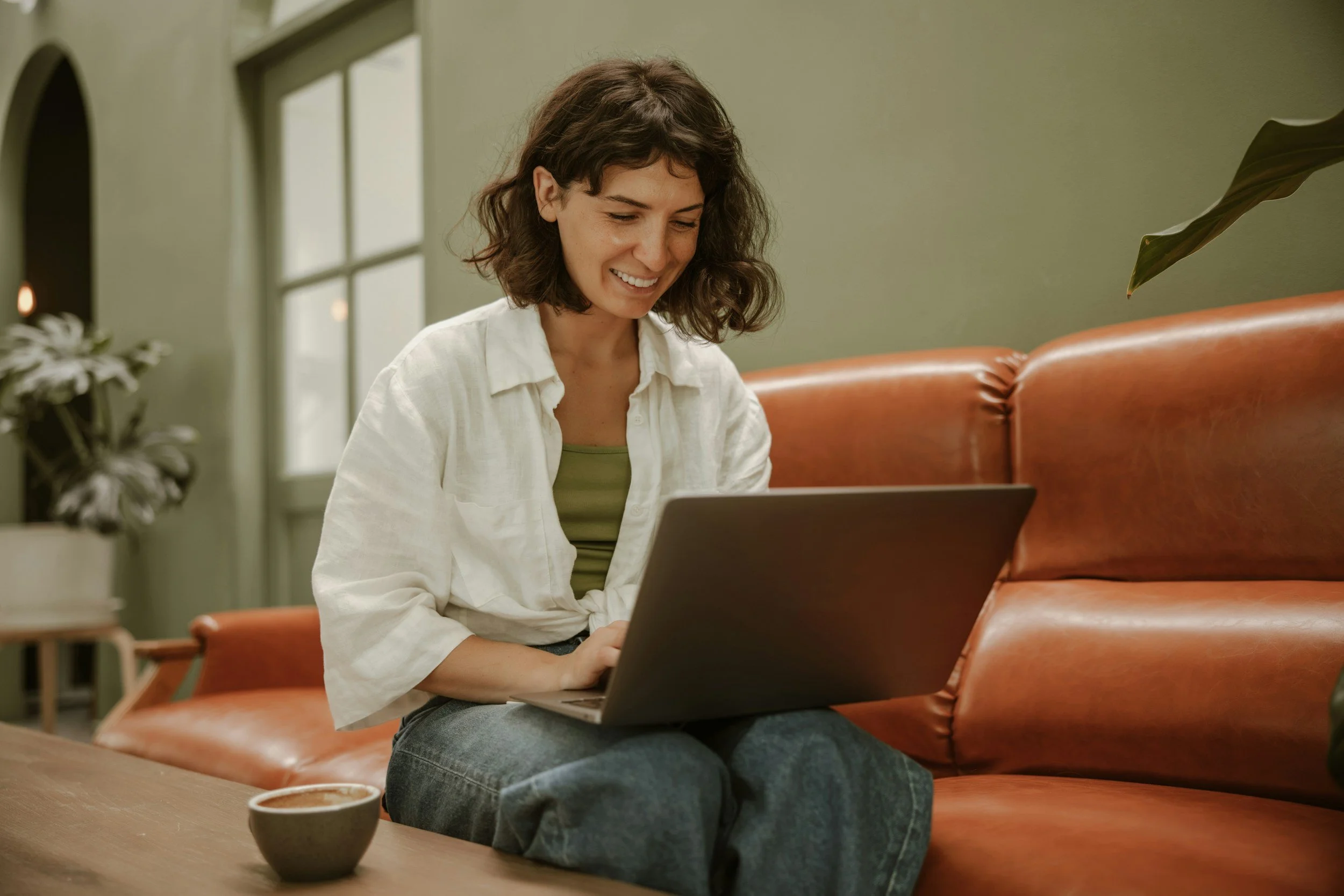Health wellness therapist smiling, sitting on brown leather couch, looking at laptop on knees with a cup of coffee on a wooden table by her side