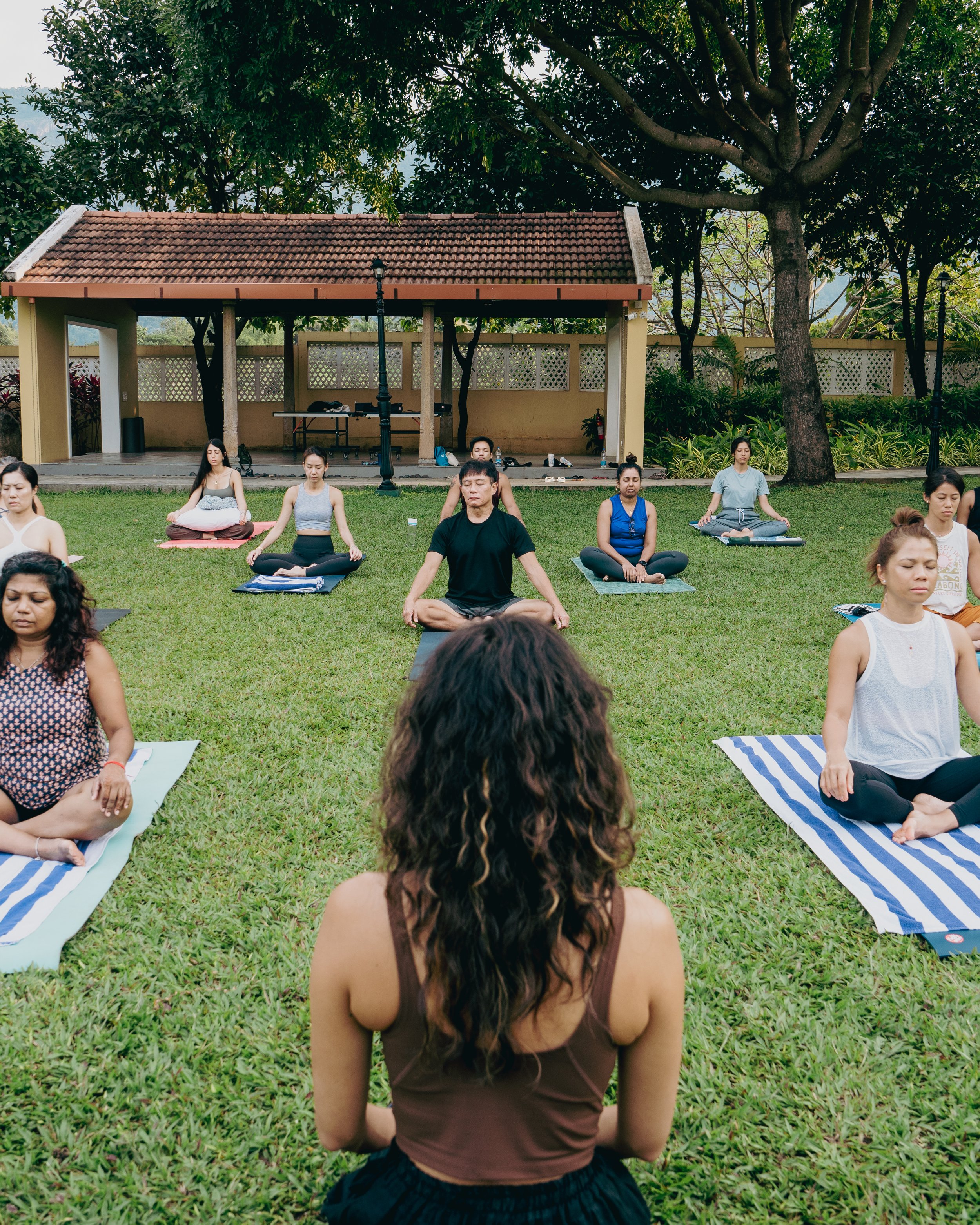 People participating in an outdoor yoga or meditation class, seated on yoga mats with legs crossed and eyes closed, surrounded by trees and greenery, with a small shelter and street lamps in the background.