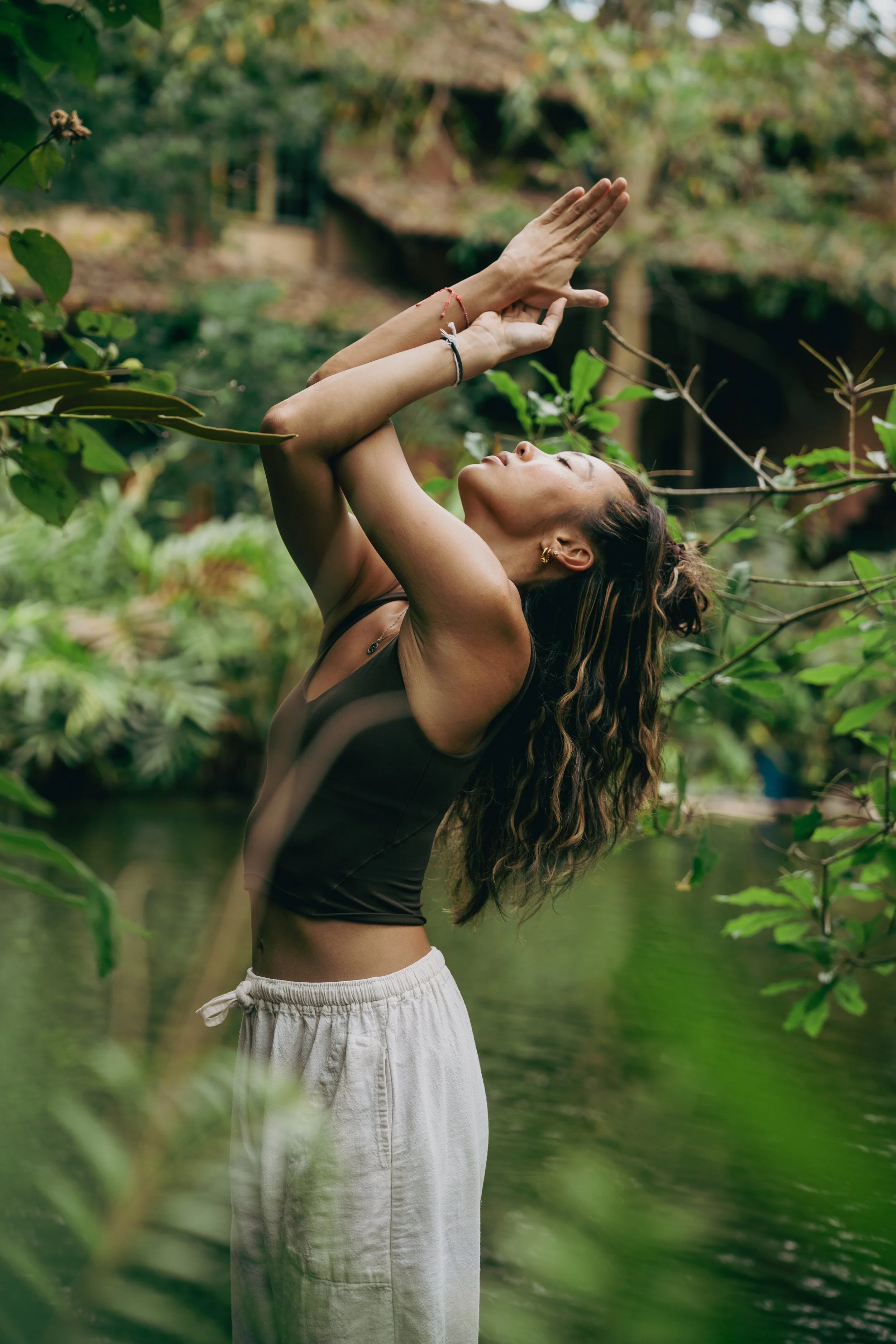 A woman standing outdoors in front of a body of water, with her arms raised above her head and facing upwards, surrounded by green trees and foliage.