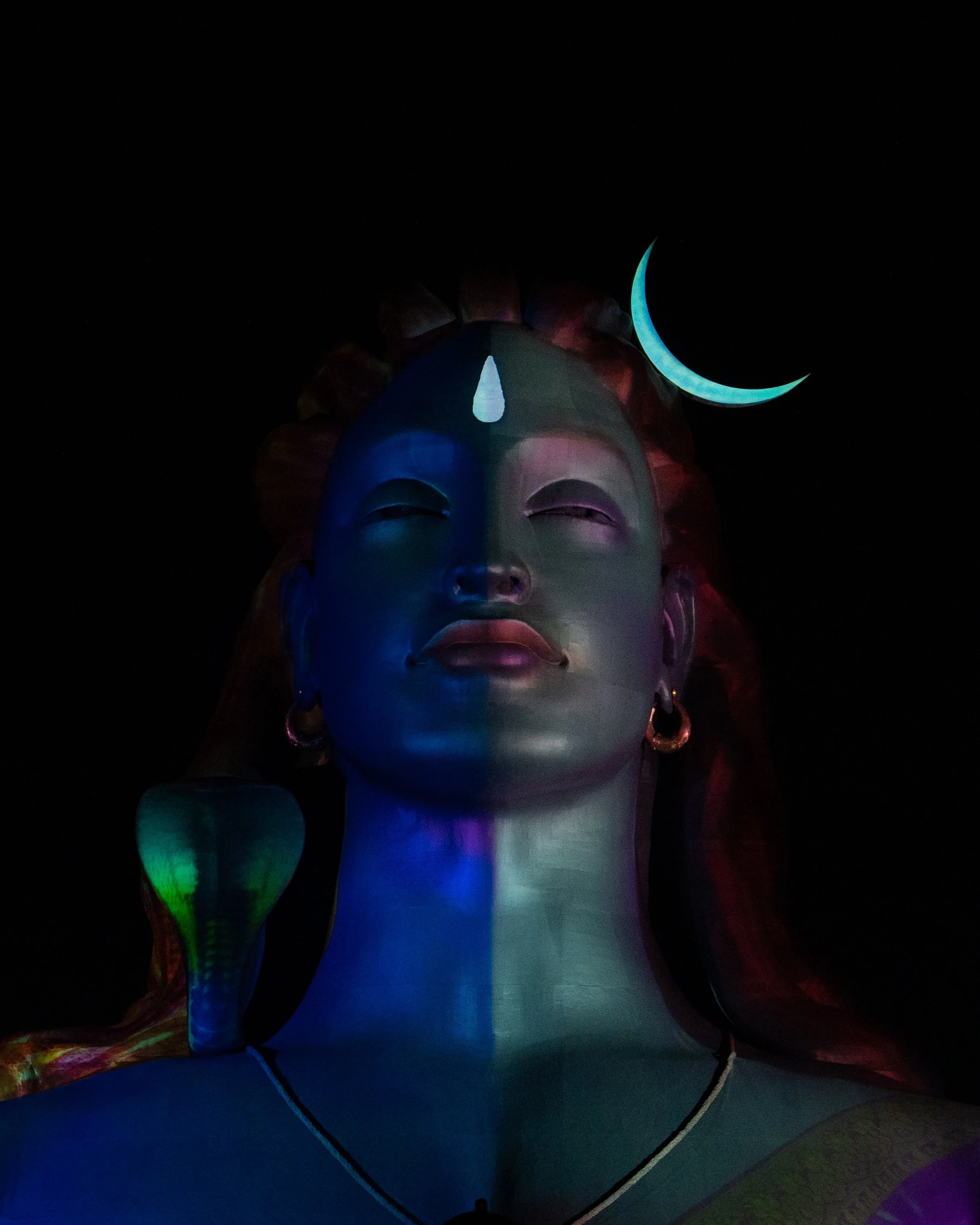 Colorful Large inflatable sculpture of a human face with closed eyes and a teardrop-shaped element on the forehead, illuminated with blue, green, purple, and red lights, set against a dark night background.