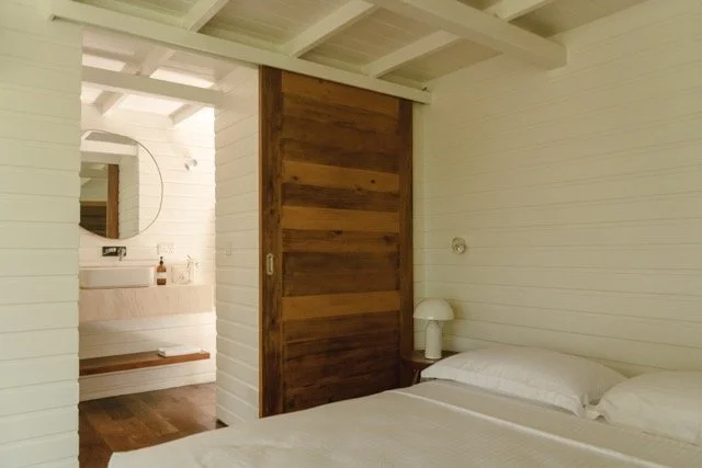 A cozy bedroom with white wood-paneled walls and ceiling, a bed with white bedding, a round mirror above a wooden vanity, and a sliding wooden door separating the sleeping area from an adjacent space.