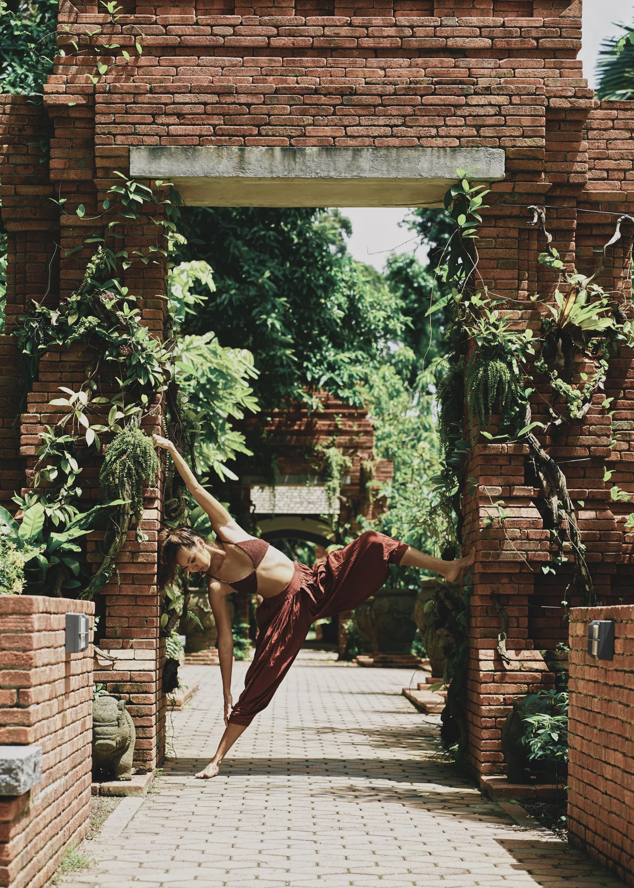 A woman practicing yoga outdoors in front of a brick archway surrounded by greenery.