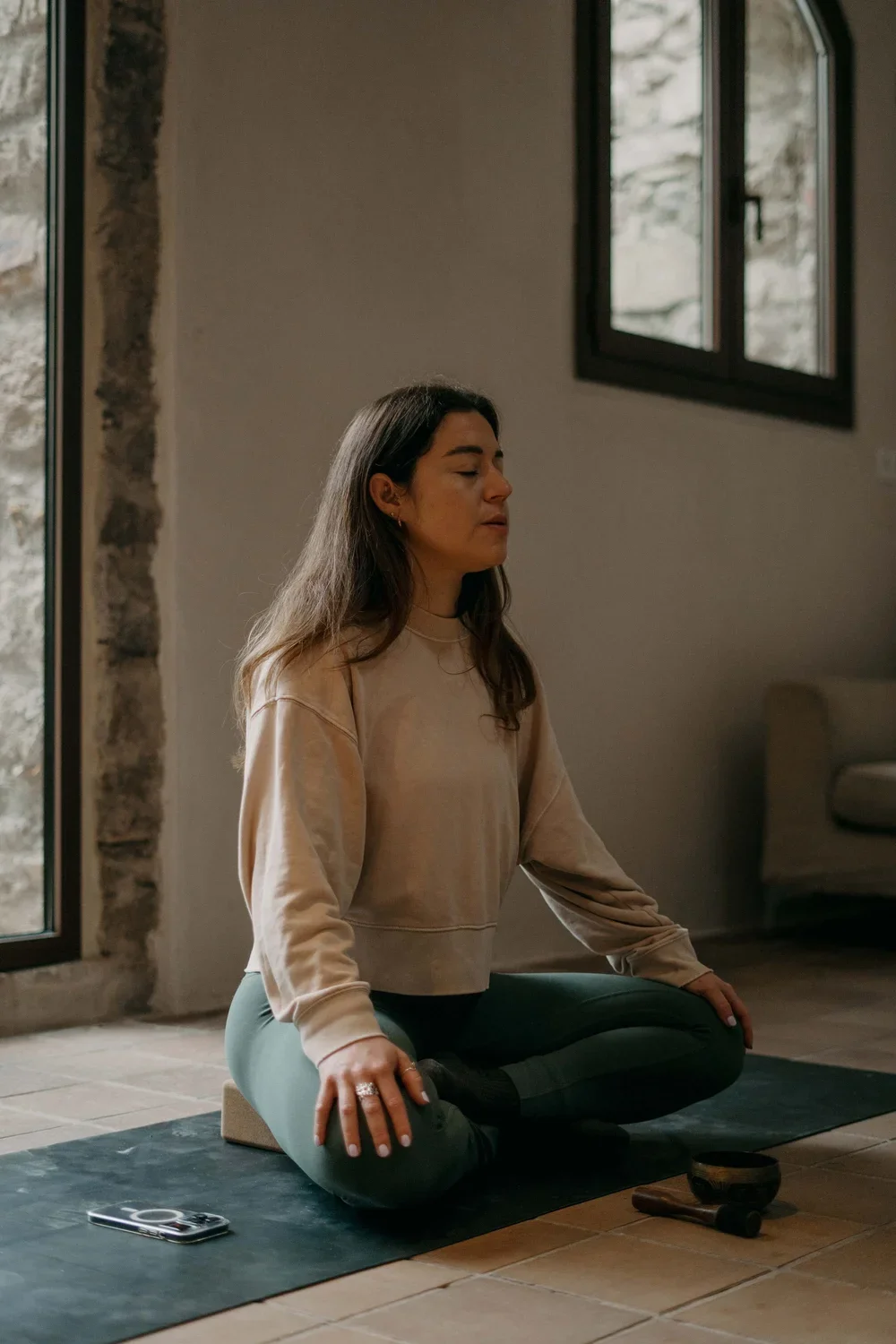 A woman practicing yoga indoors, sitting cross-legged on a mat, with closed eyes and a calm expression, near a window with natural light.
