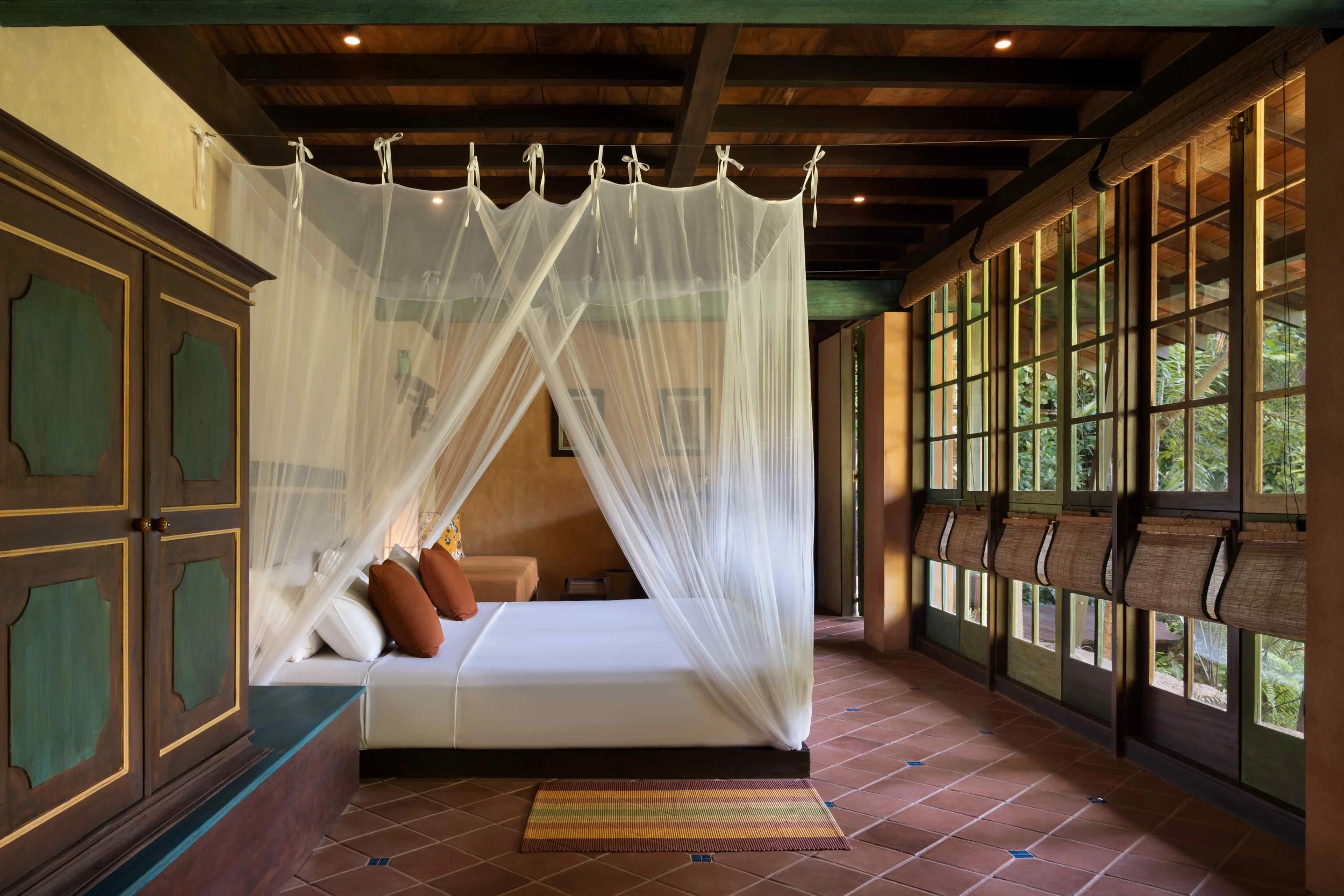 A bedroom with a four-poster bed with sheer white curtains, wooden ceiling, large windows with woven blinds, and a colorful striped rug.