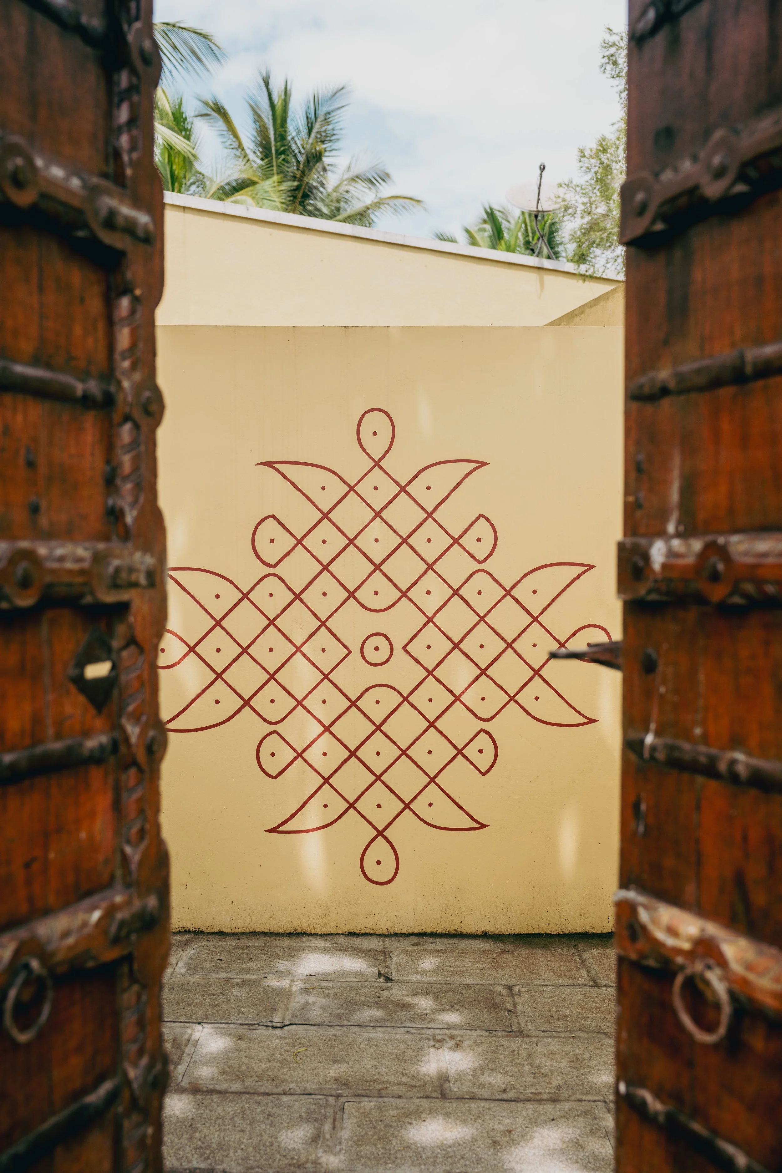 Open wooden door framing a view of a decorative wall with a symmetrical red ornament, a yellow wall, and green trees with palm fronds in the background.