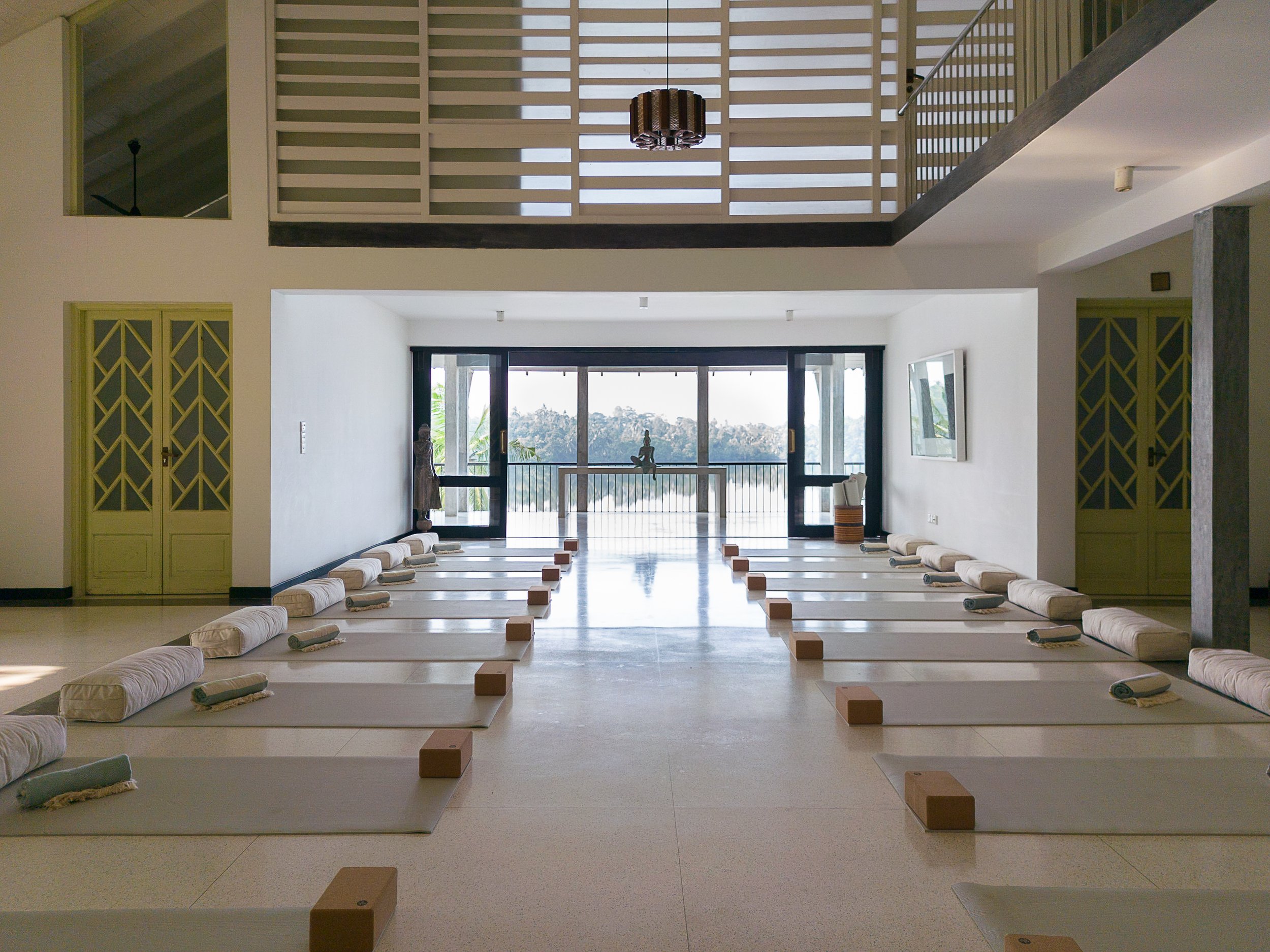 A spacious room with short white cushions and yoga mats arranged on the floor, set up for meditation or yoga practice, with large sliding glass doors leading outside to a scenic view.