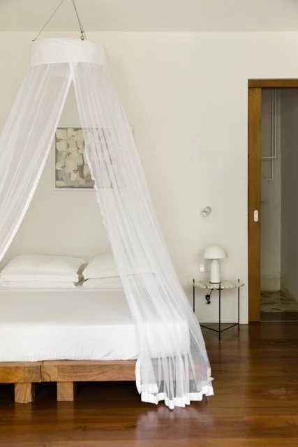 A bedroom with a white bed and a canopy with sheer white curtains, a nightstand with a white lamp, and a wooden sliding door.