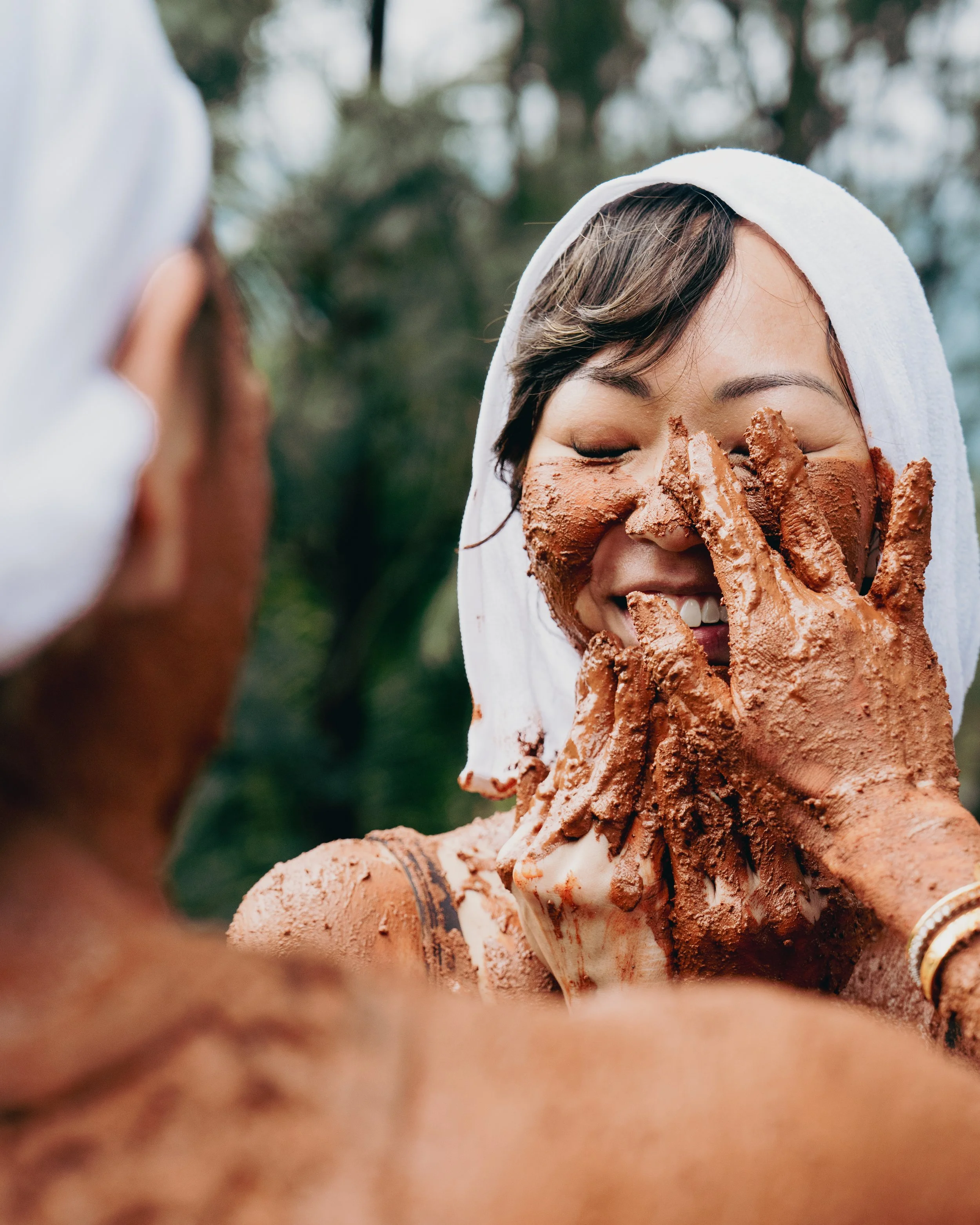 A woman with closed eyes, wearing a white towel on her head, holding a large chocolate ice cream cone covered with chocolate and cookie pieces, outdoors with blurred trees in the background.