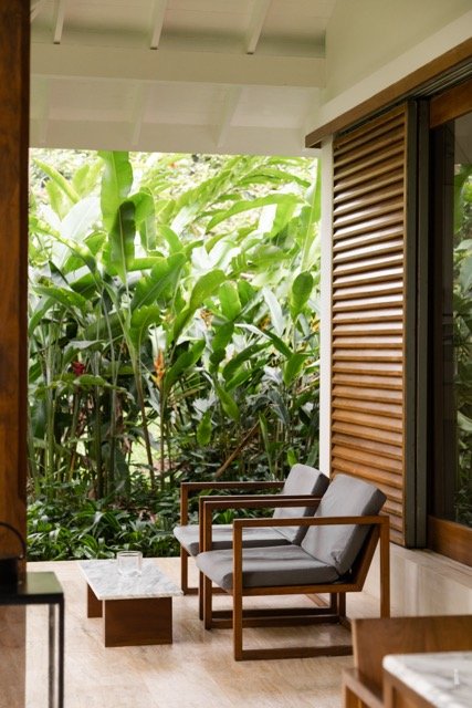 Patio area with two wooden chairs with gray cushions and a small coffee table, overlooking lush green jungle foliage outside a house with wooden shutters.