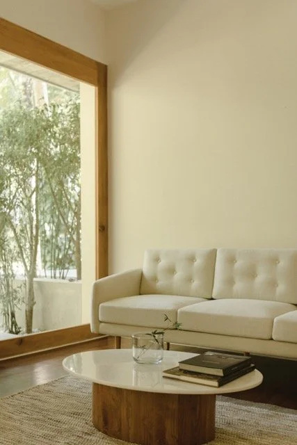 Modern living room with a white tufted sofa, wooden coffee table with books, glass vase, large window with a wooden frame, and view of trees outside.
