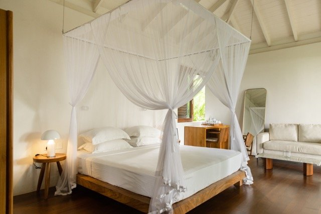 Bedroom with a four-poster bed draped with white canopy, white bedding, wooden side table with lamp, wooden vanity with mirror, and white sofa in the background.