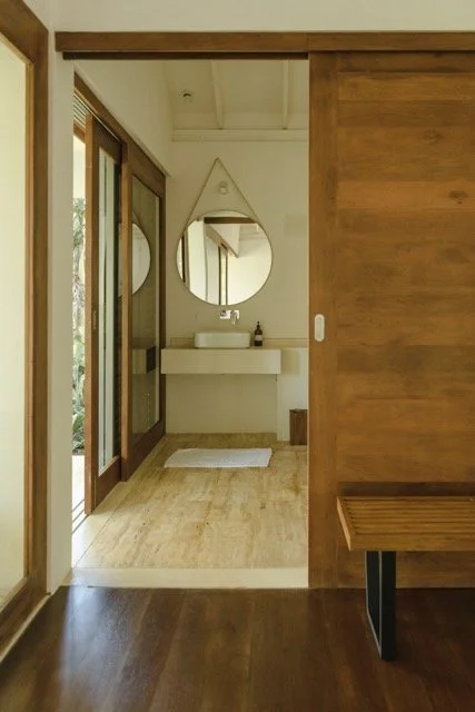 Bathroom with circular mirror above a white sink, wooden bench beside it, and sliding wooden door in a modern, minimalist style.