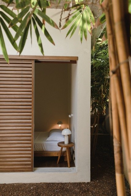 Interior view of a bedroom seen through a wooden louver door with greenery outside.