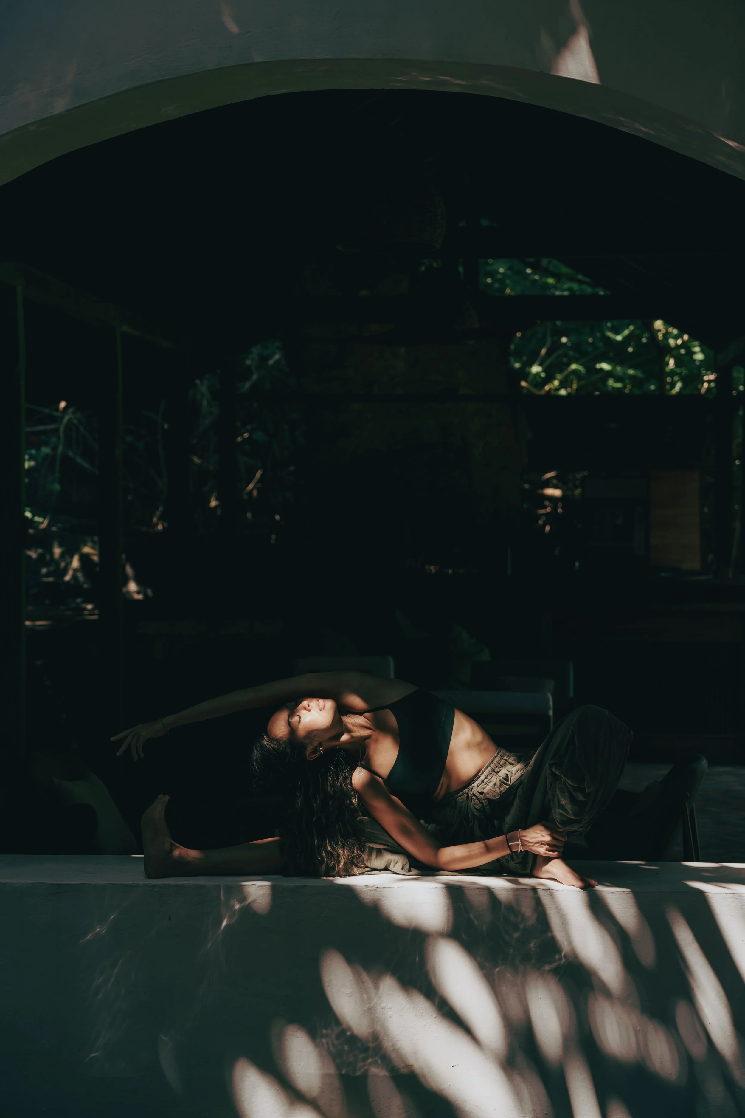 A woman practicing yoga indoors, sitting on the floor in a side stretch pose with her eyes closed, wearing a black sports bra and beige pants. Natural light coming through the windows, with plants and furniture in the background.
