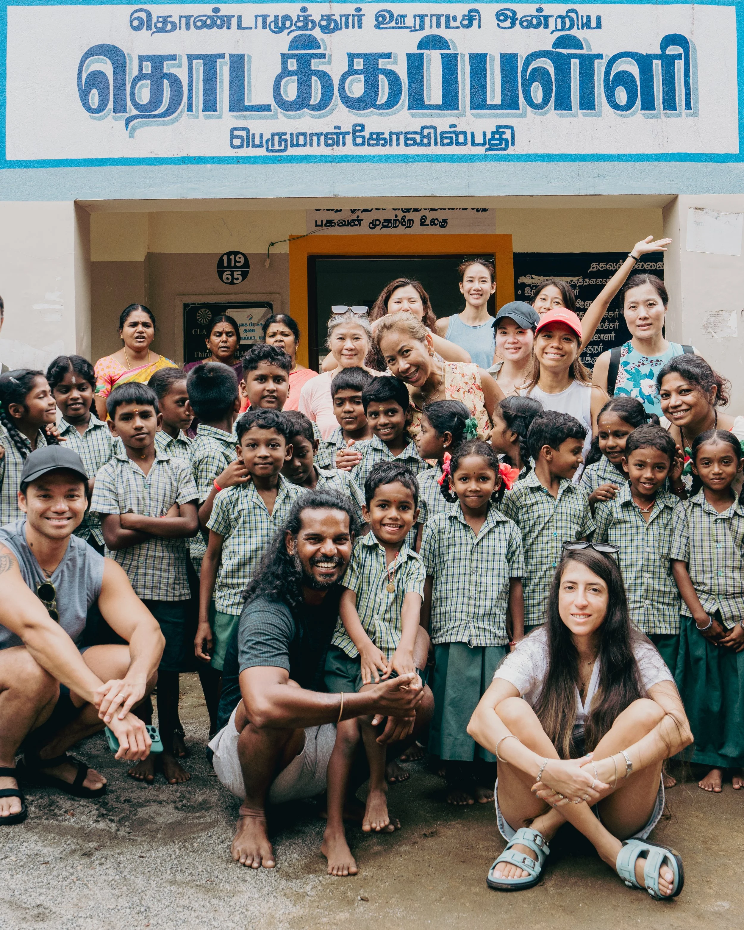 A group of children and adults standing outside a school building with Tamil signage, smiling and posing for a photo.