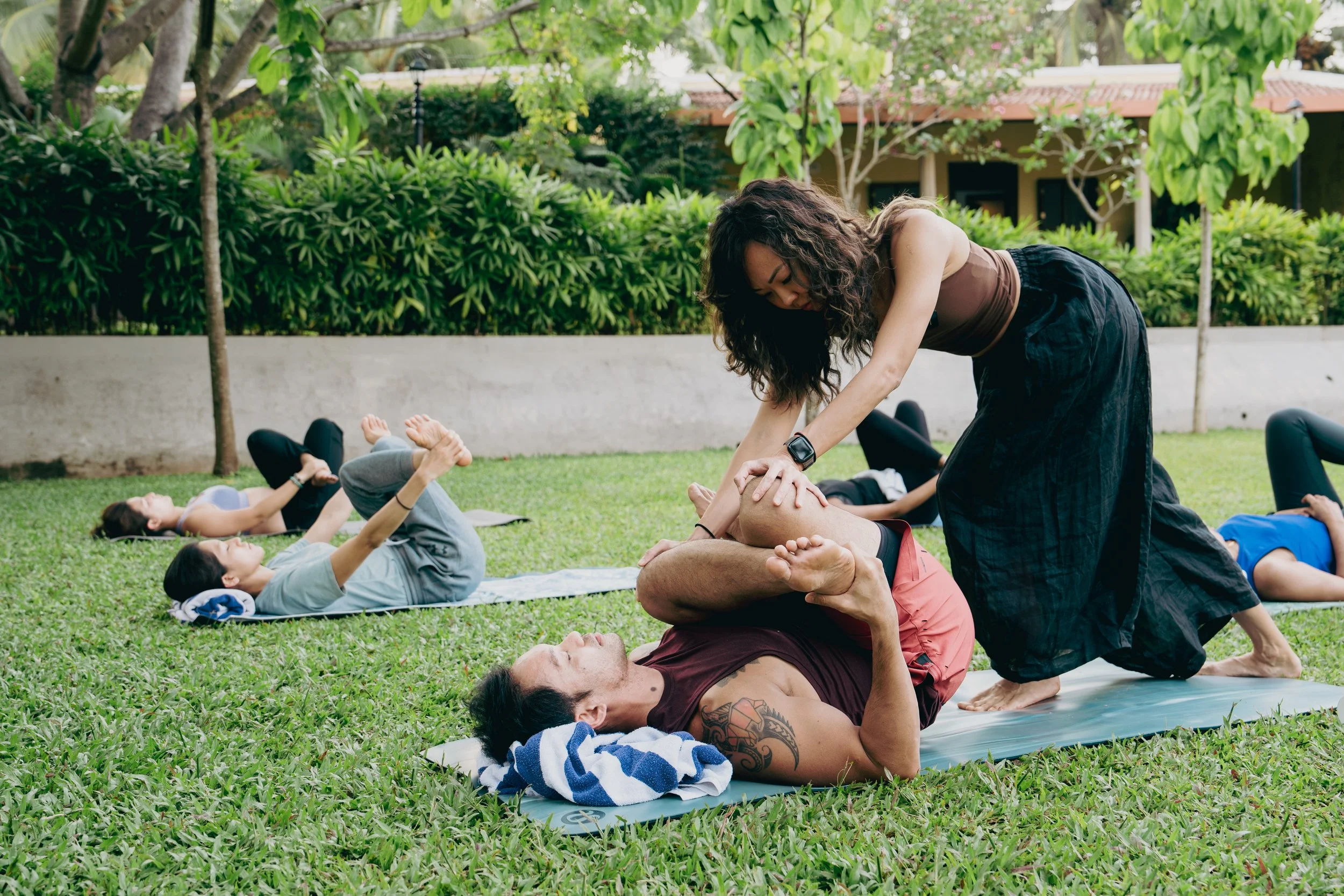 A group of people participating in an outdoor yoga or fitness class on a grassy field, with a woman assisting an older man with a stretch or pose, in a park with trees and a building in the background.