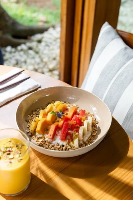 A bowl of cereal topped with sliced mango, strawberries, bananas, and edible flowers on a wooden table with a knife and fork nearby, sunlight casting shadows inside a cozy dining area.