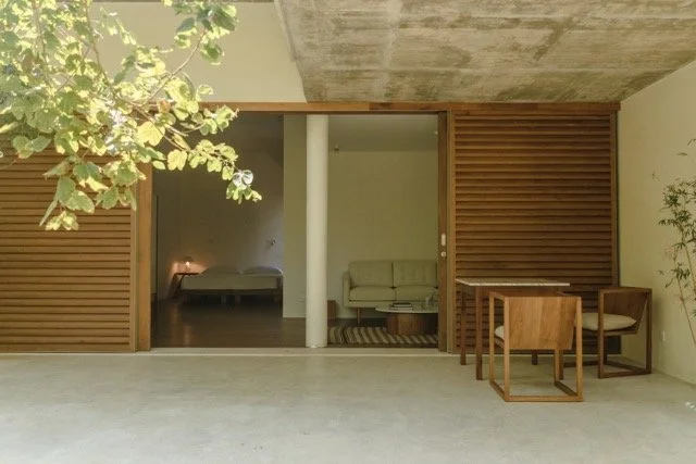 View of a modern living space with wooden paneling, sliding doors, a sofa, small dining table with chairs, and a bed in the background.